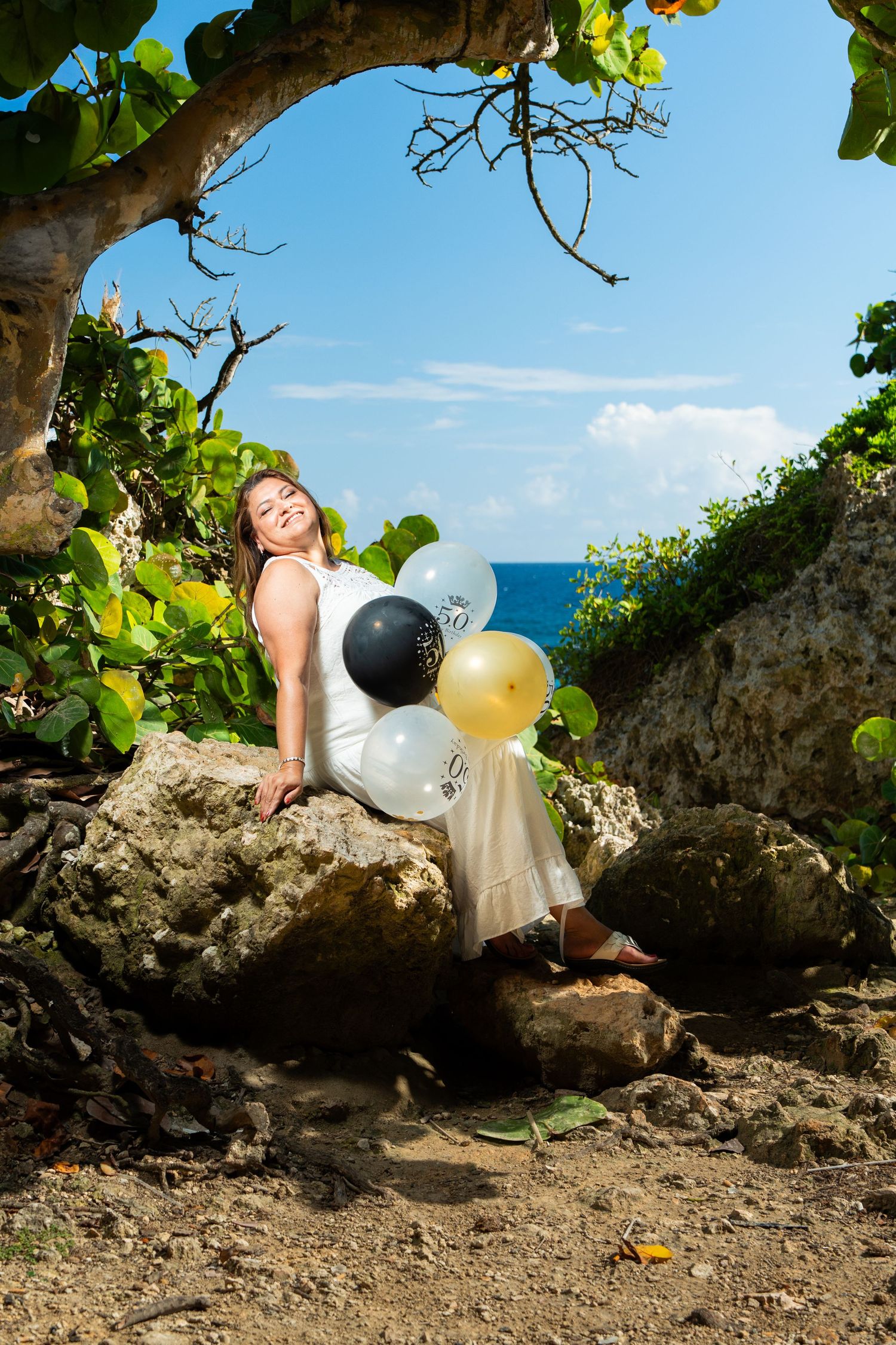 Young woman in white dress holds colorful balloons while sitting on rocks near tropical ocean coastline.