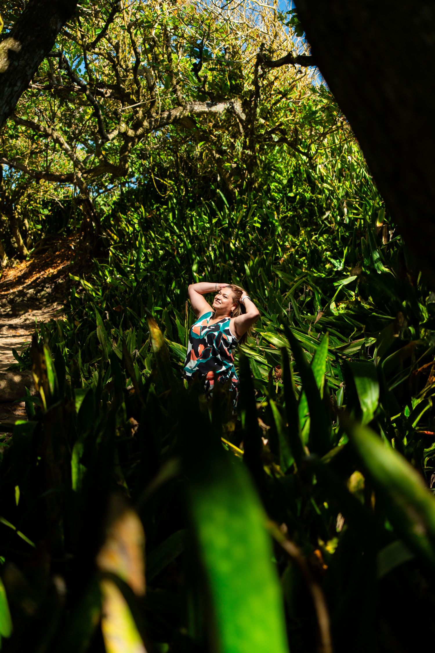 Woman in colorful floral dress stands among lush green tropical foliage in sunlit forest setting.