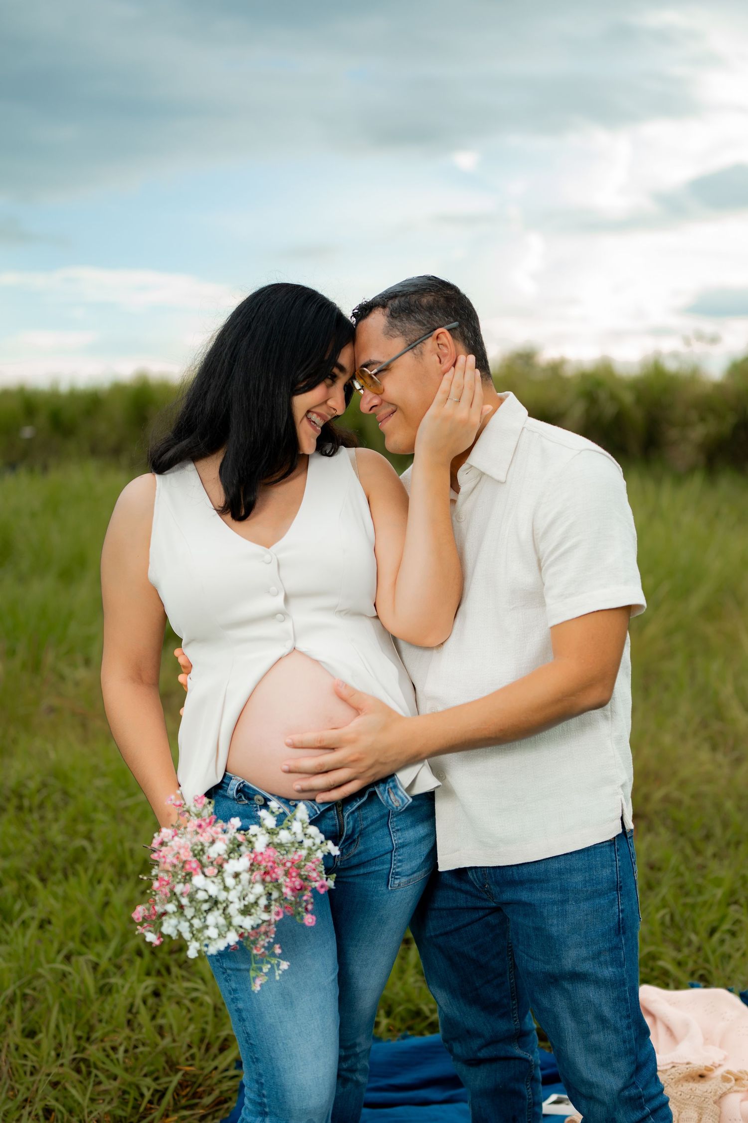 Happy pregnant couple embracing in green field holding wildflowers under cloudy sky.