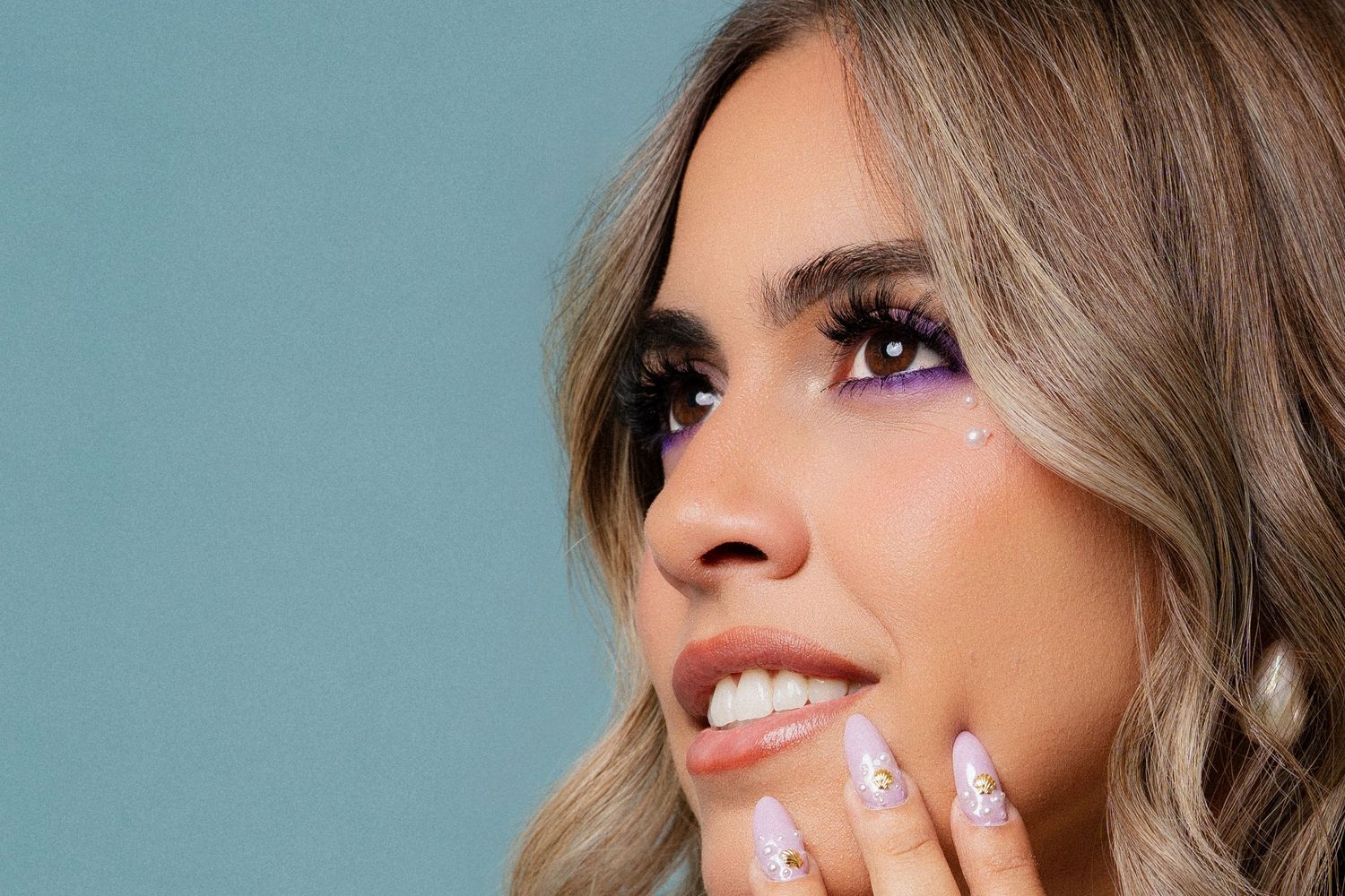 Close-up of woman with purple glitter eye makeup and decorated nails against blue background.