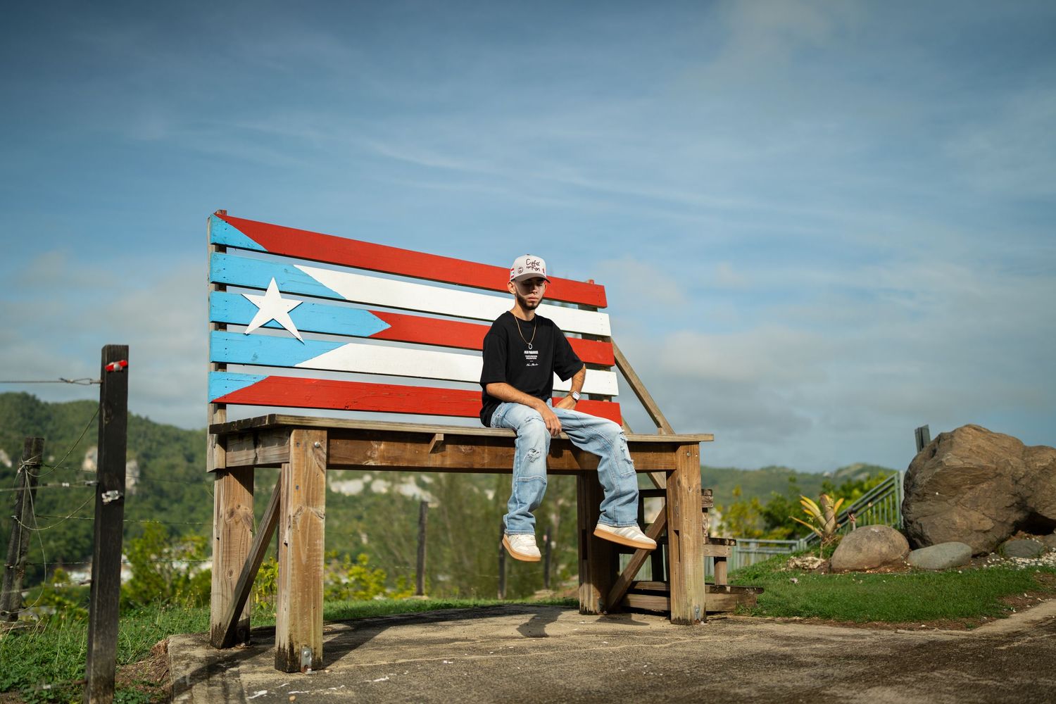Man sitting on a large Puerto Rico flag painted wooden bench outdoors under a blue sky.
