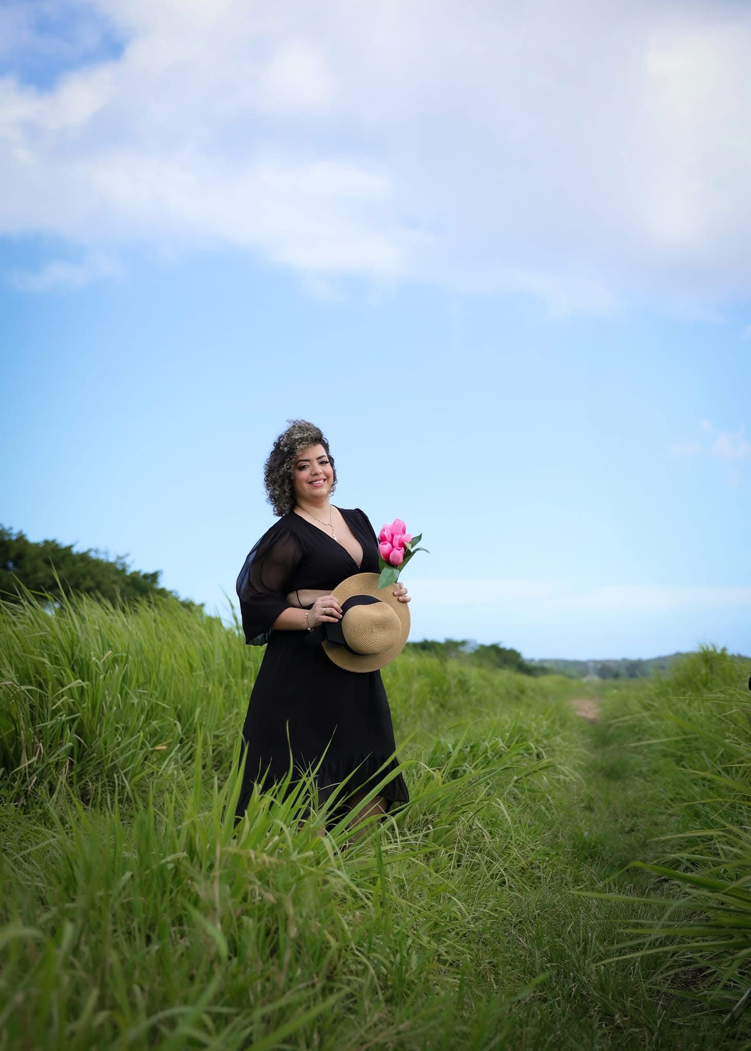 Plus-size woman in black dress holding pink flowers and straw hat in meadow.