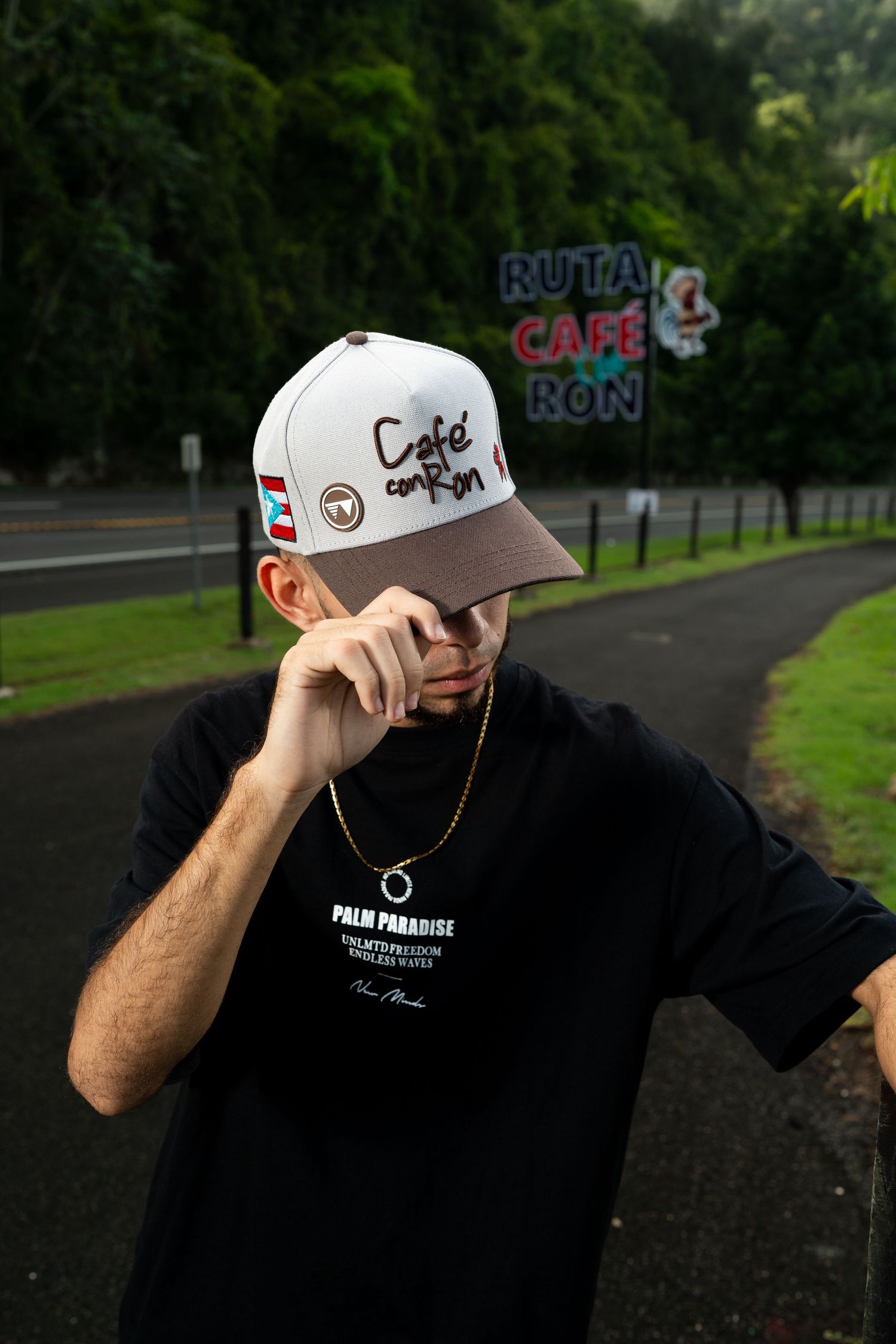 Man in black shirt wearing Cafe con Ron cap poses on road with neon sign in background.