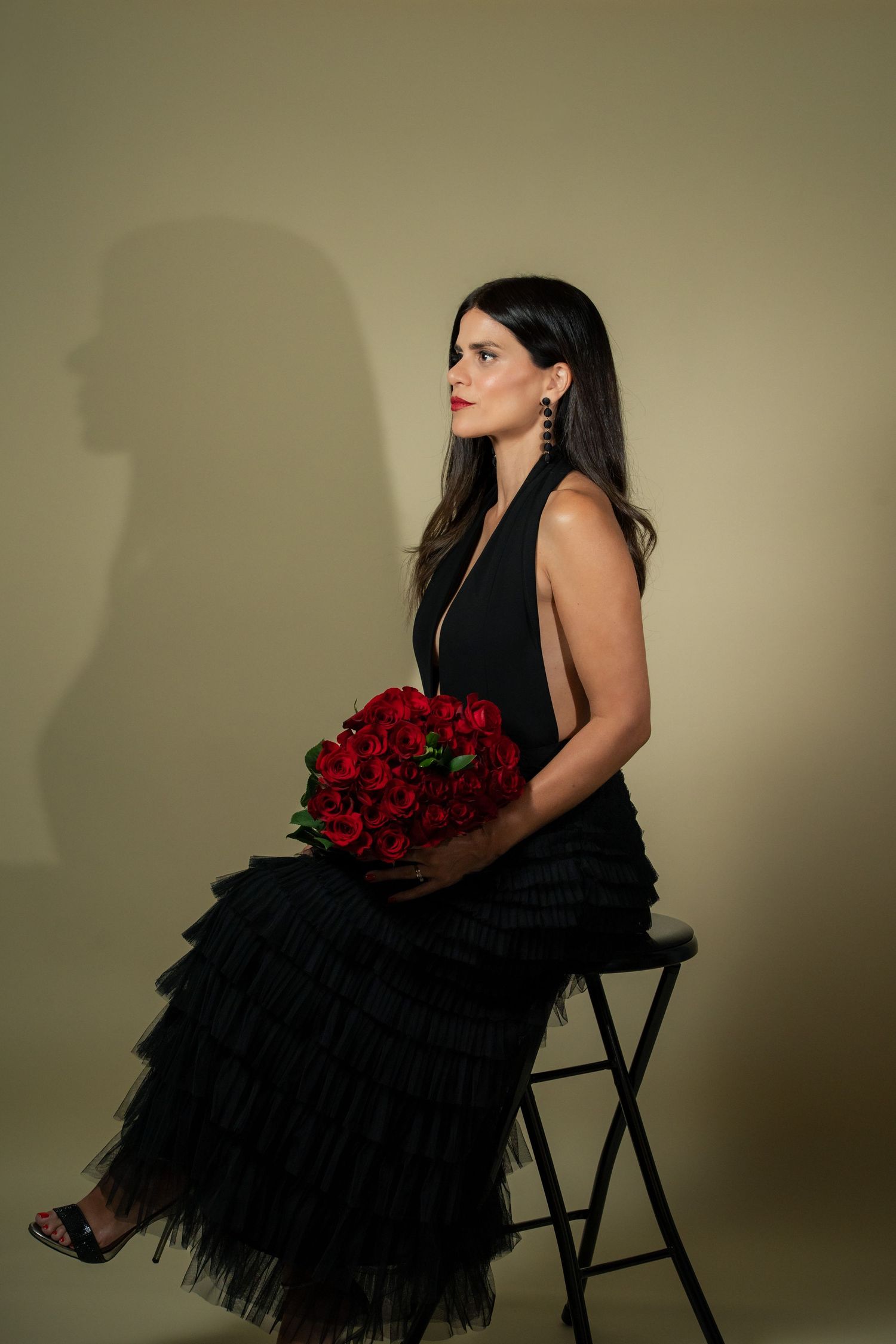 Elegant brunette woman in black dress holding red roses bouquet seated on stool.