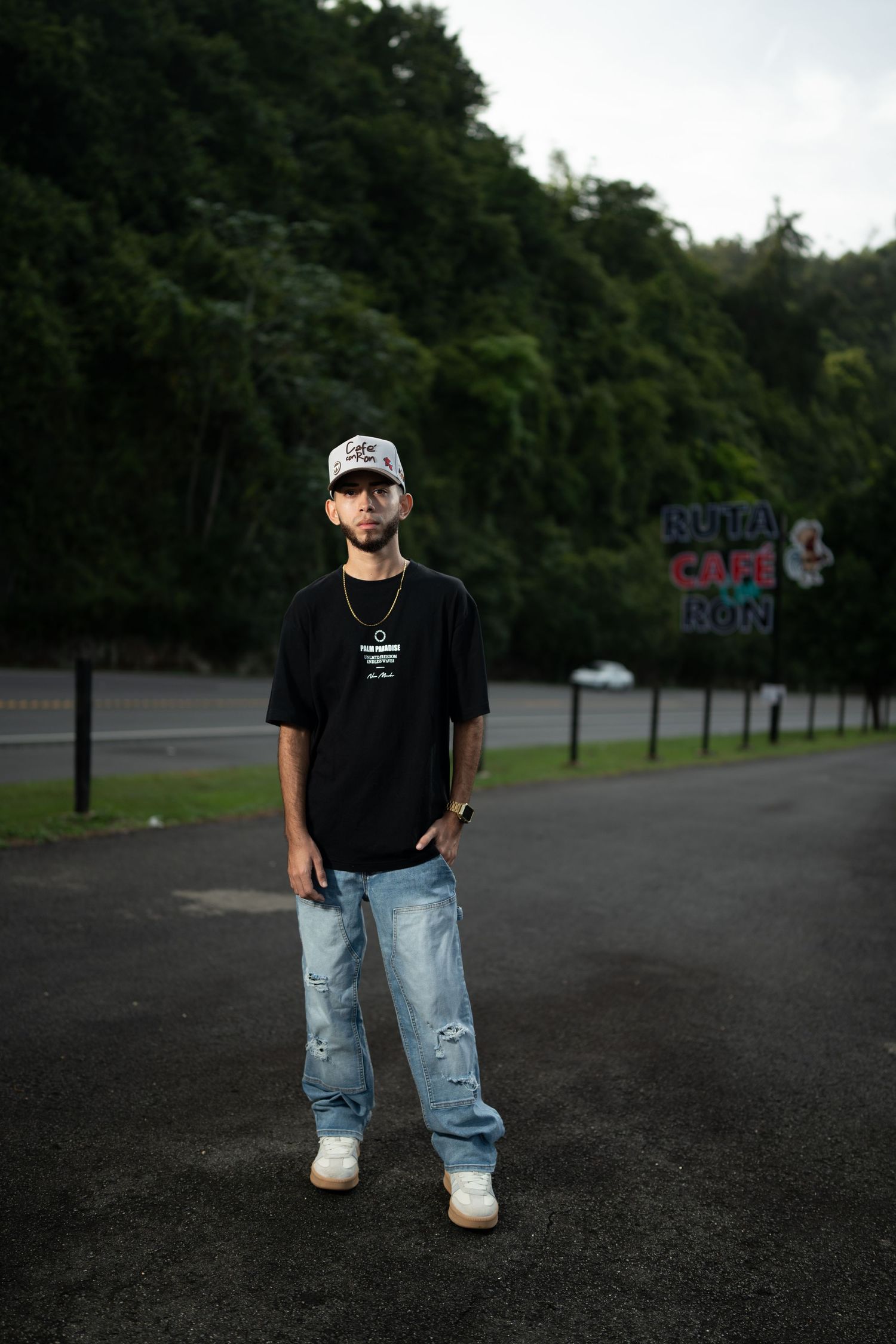 Young man in black tee, ripped jeans, and cap standing in a parking lot with green trees behind.
