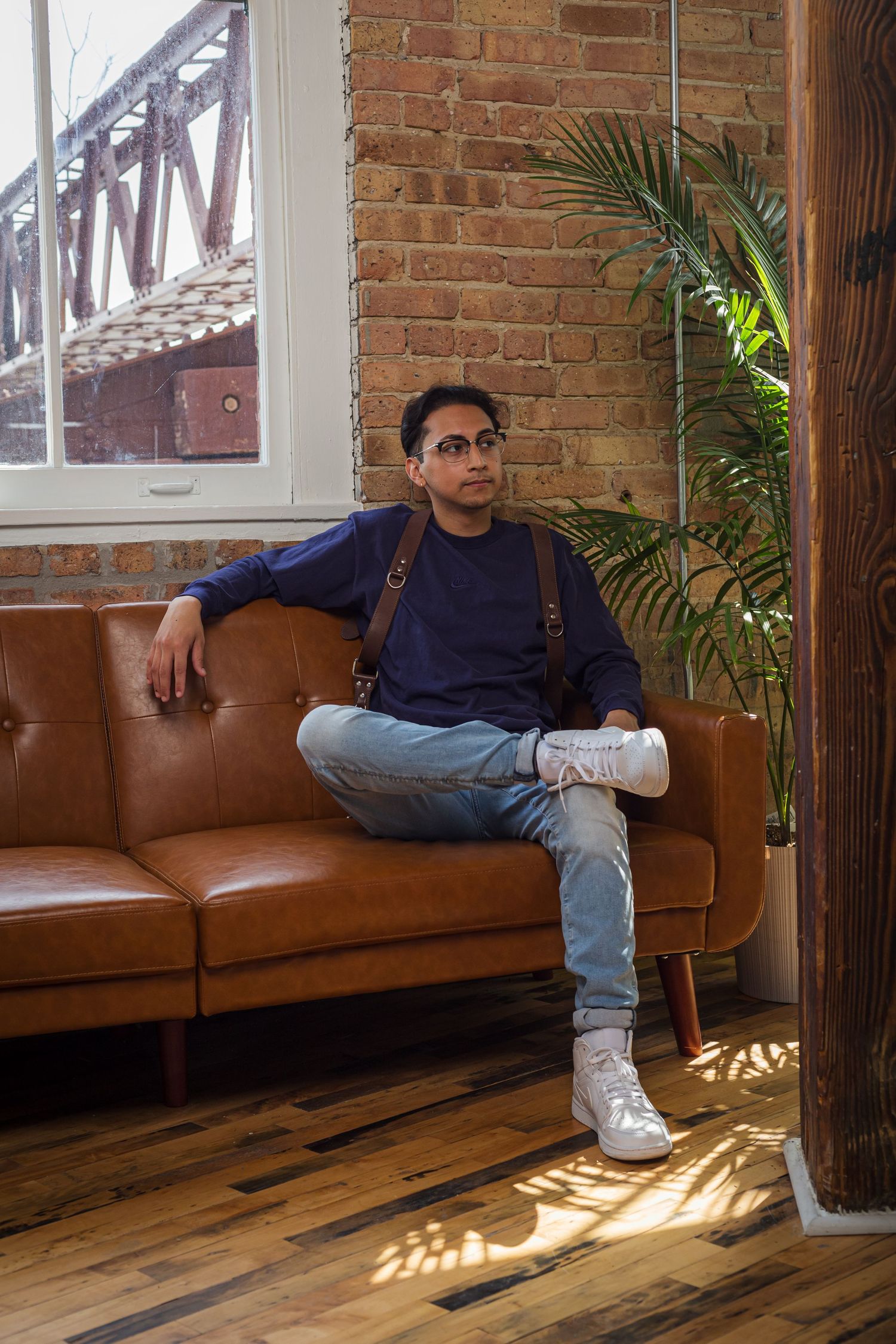A person relaxes on a brown leather couch next to a palm plant in an industrial-style room with exposed brick walls.