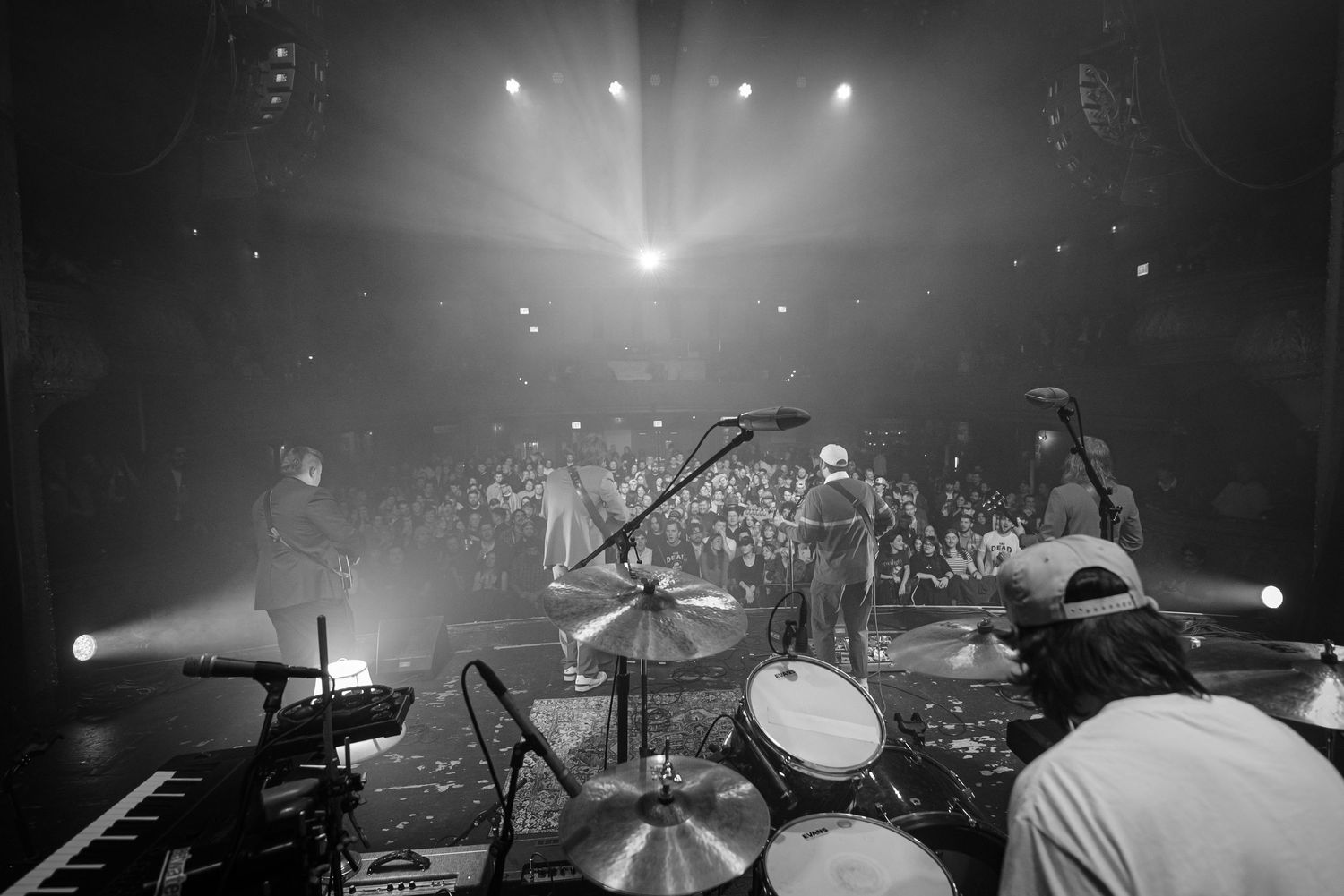 A black and white concert photo showing a stage with bright spotlights and silhouettes of performers through atmospheric haze.