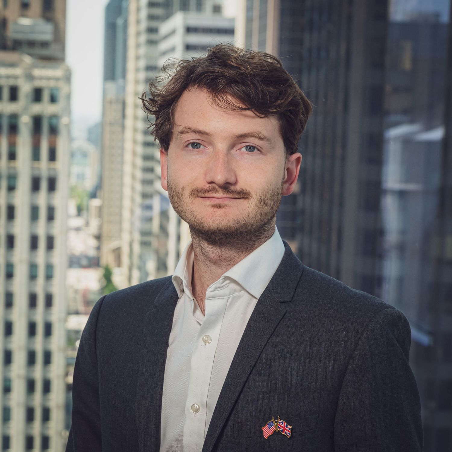 Professional headshot of someone in a white shirt and dark suit jacket against an urban office building backdrop.