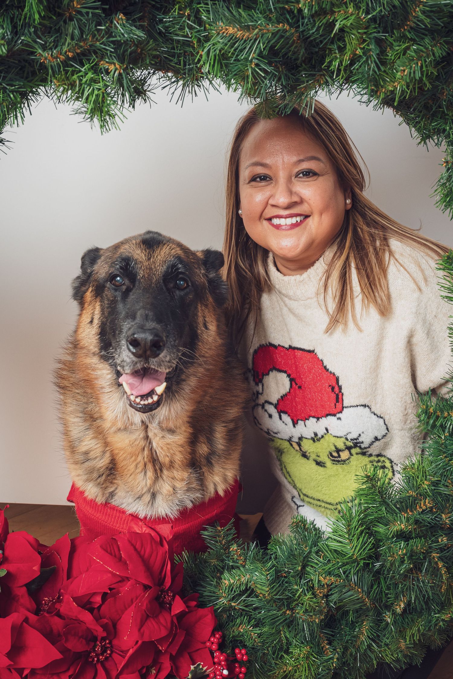 A German Shepherd wearing a red scarf poses with someone in a festive sweater next to Christmas decorations. goose.img photography LLC
