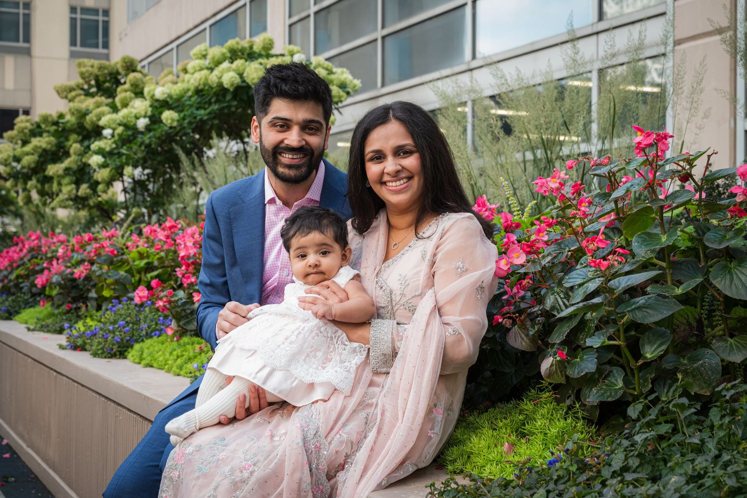A family poses together outdoors on steps near blooming pink flowers and greenery in coordinated light-colored outfits.