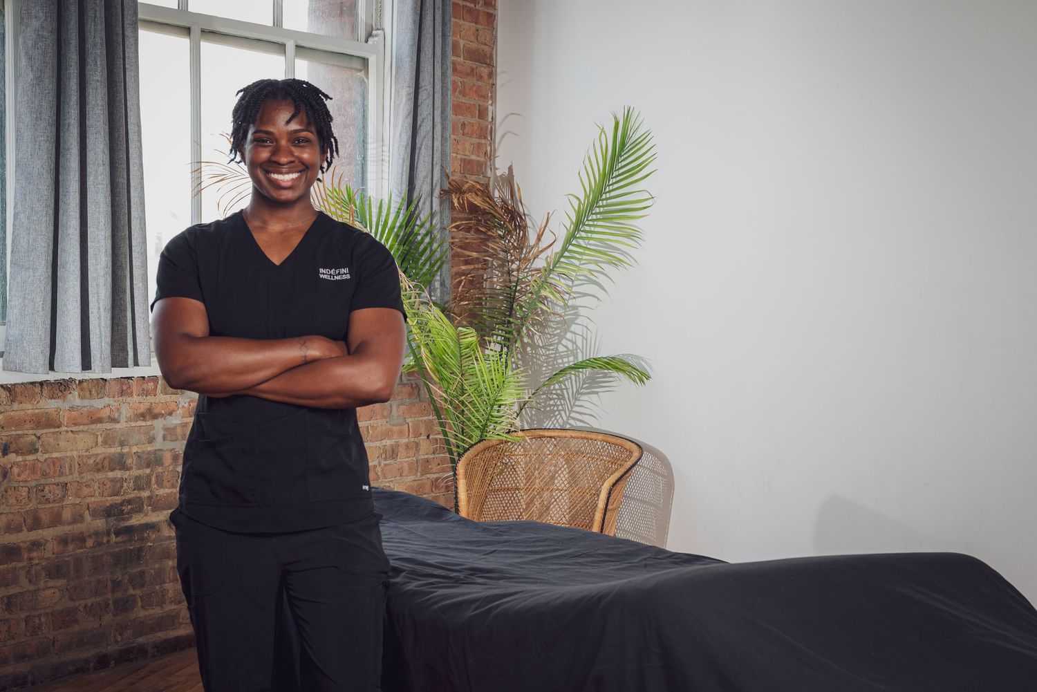 A massage therapist in black attire stands with crossed arms next to a treatment table and palm plant.