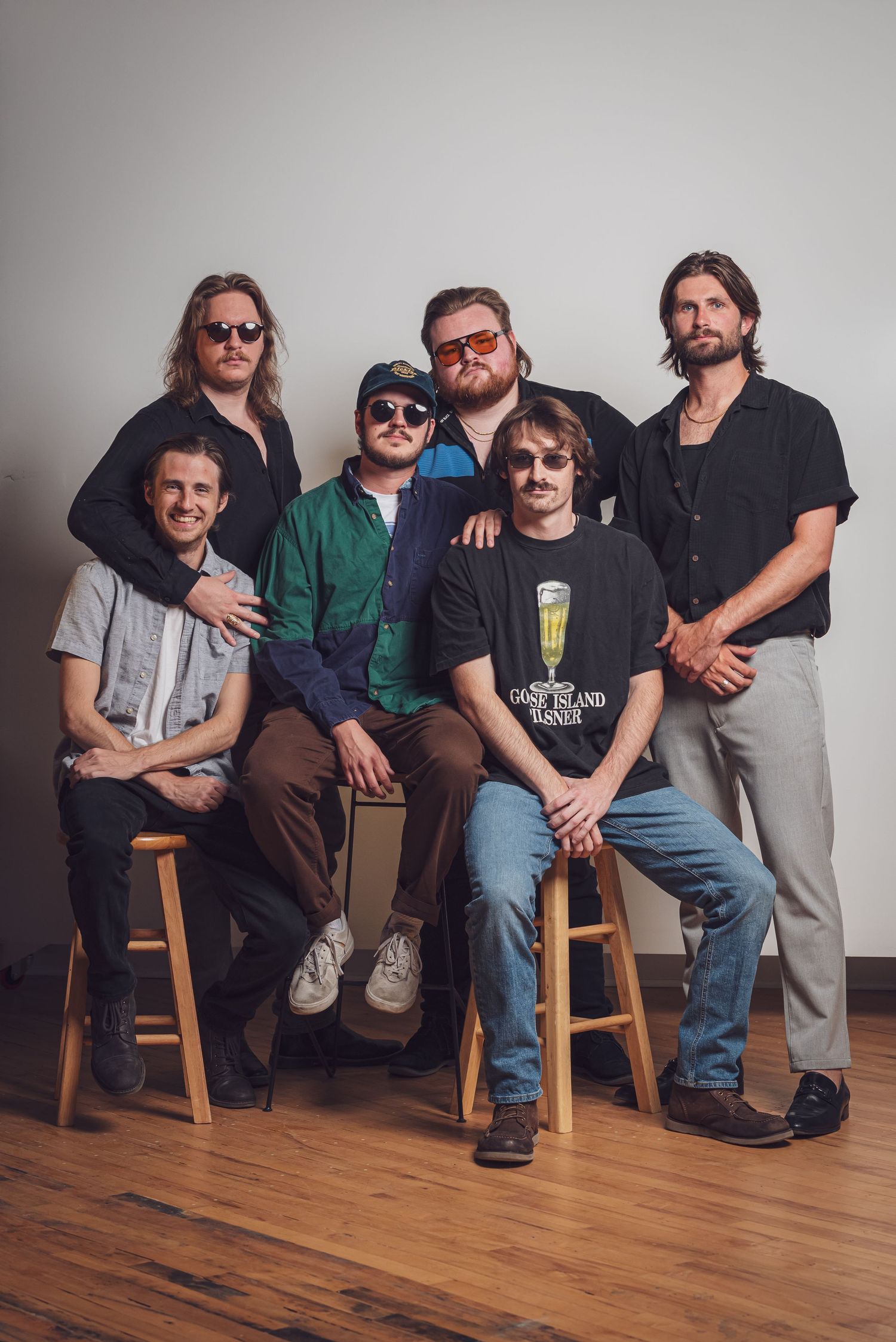 Five musicians sitting on wooden stools against a white wall in a casual group photo series.