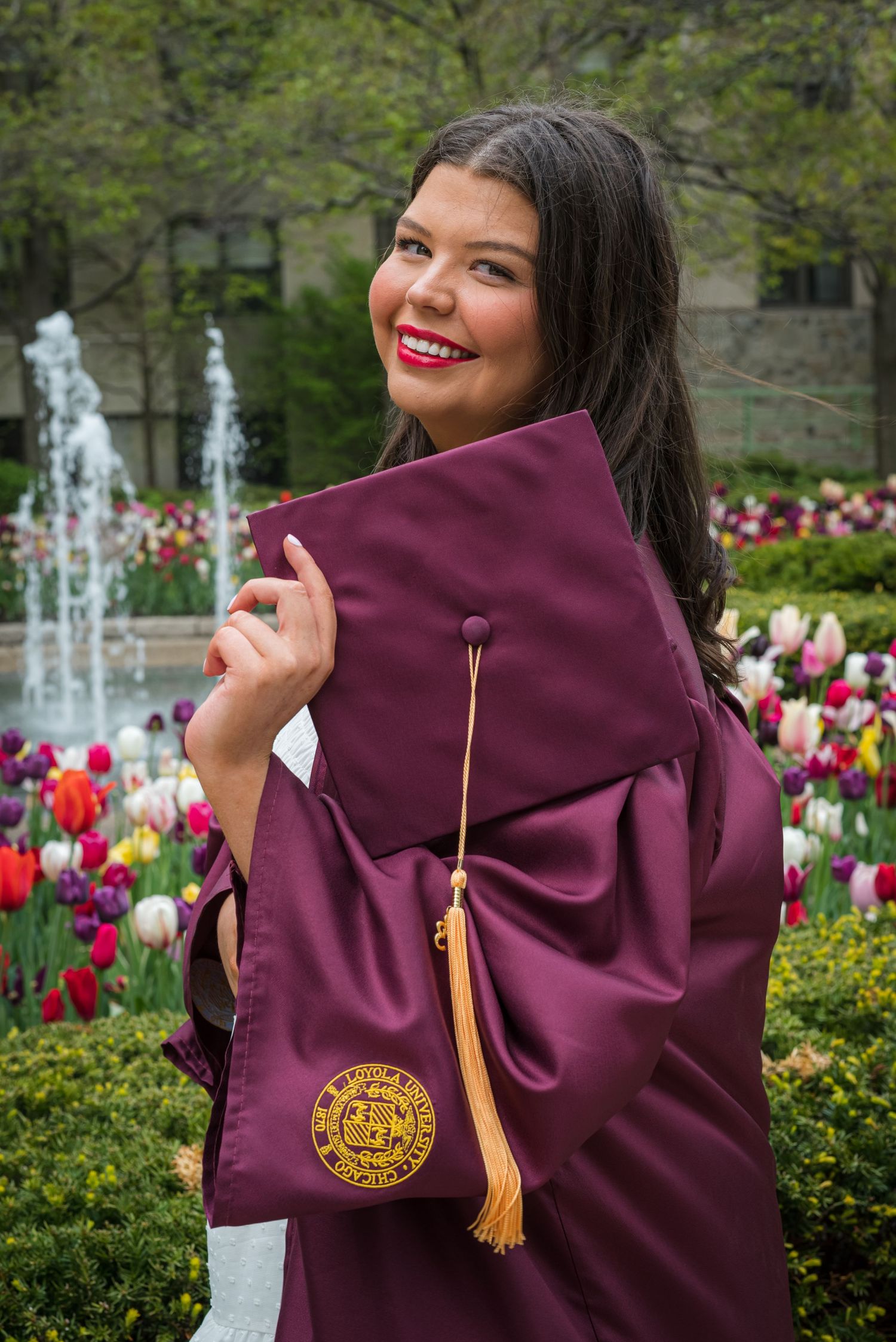 A graduate in maroon regalia poses in a garden with blooming tulips and a fountain. goose.img photography LLC - Chicago Photographer