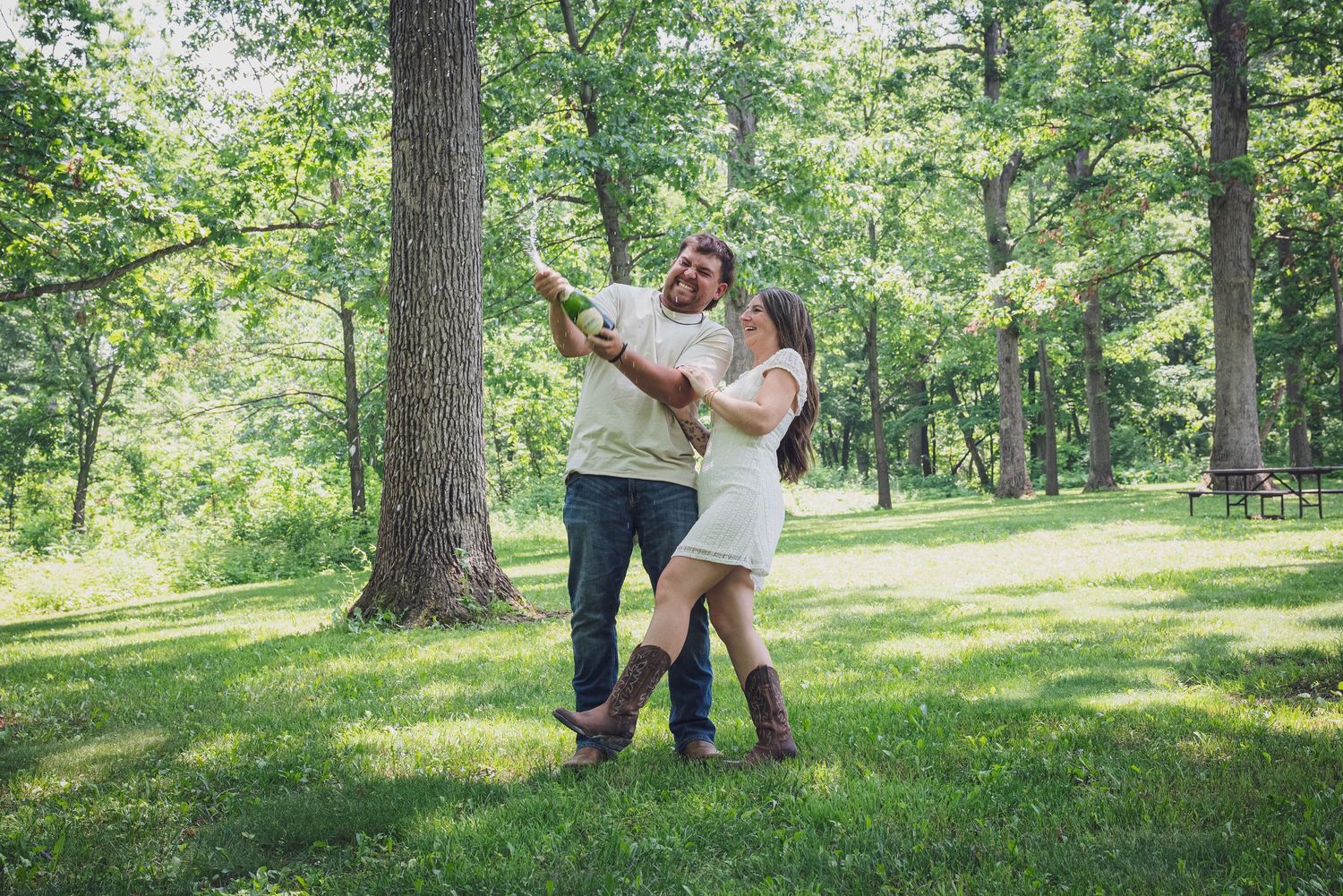A playful couple dances together on green grass in a sunlit park setting.