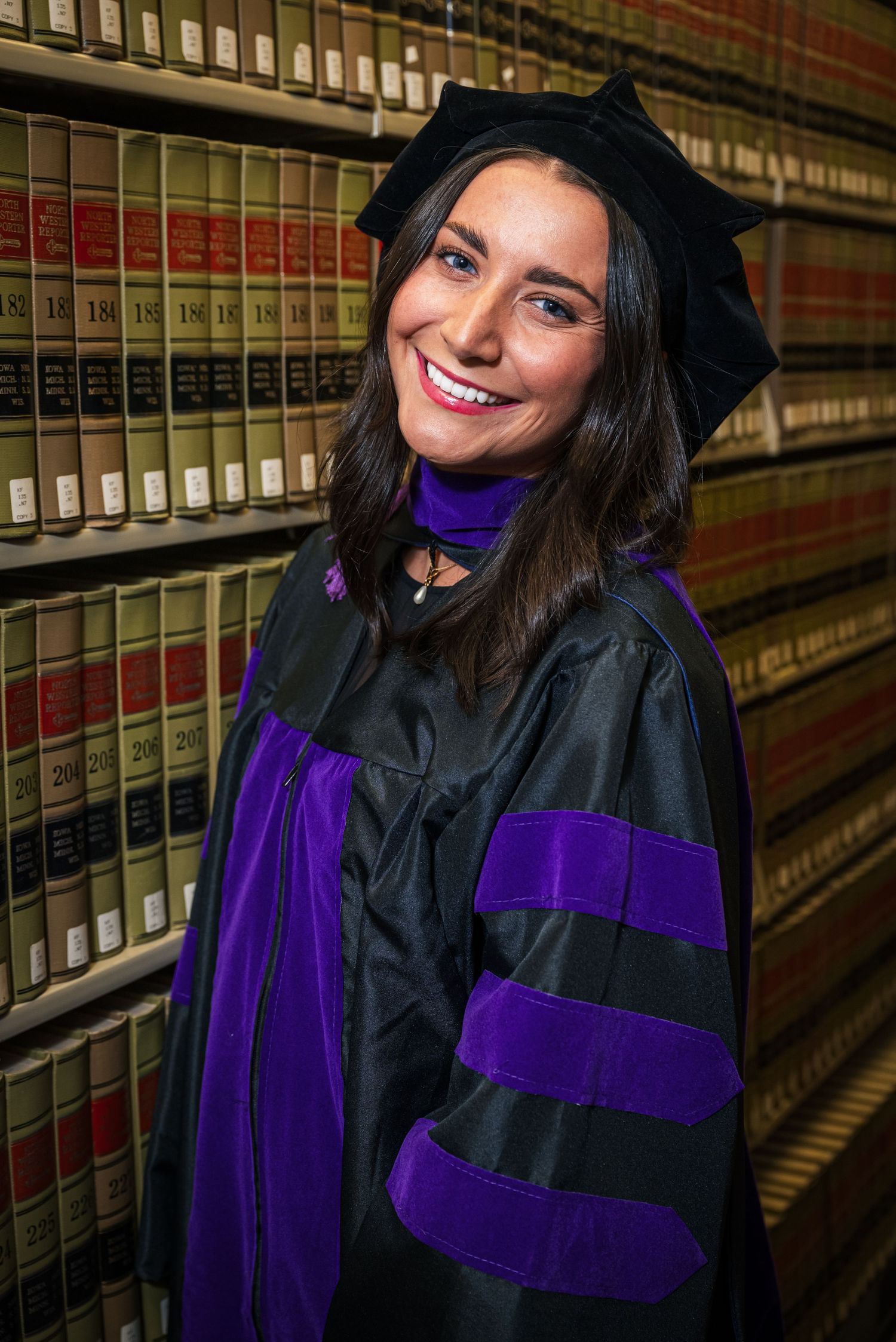 Graduate in black and purple doctoral regalia smiles while standing in front of law library bookshelves.