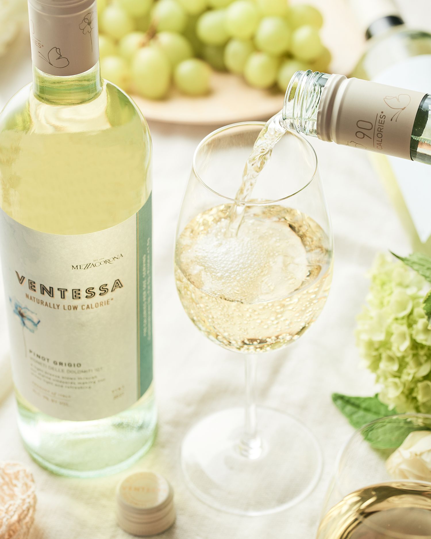 White wine being poured into a crystal glass with grapes and green hydrangeas in the background.
