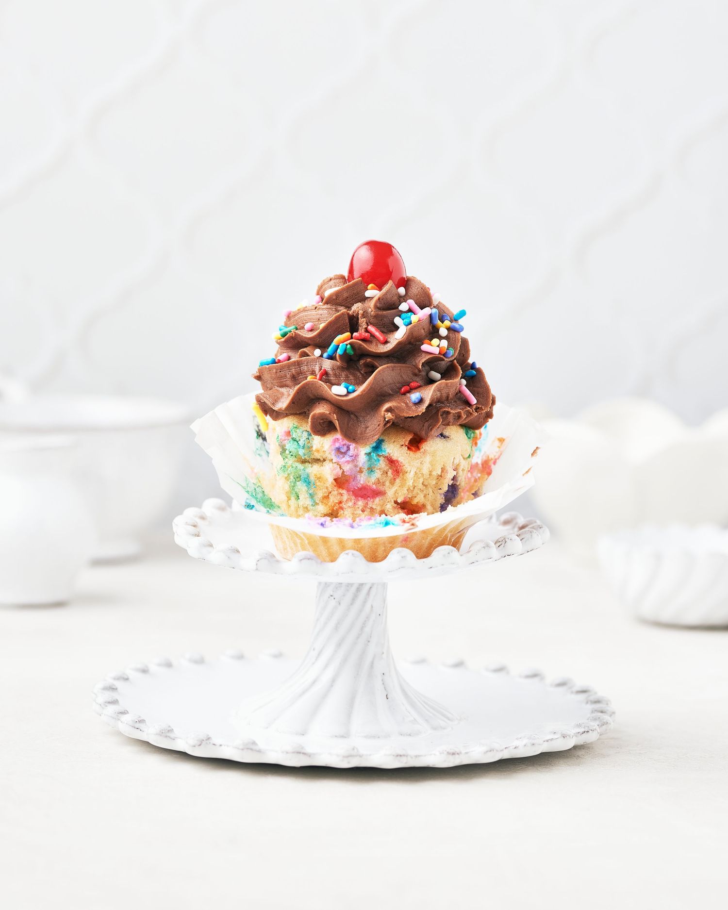 Chocolate cupcake with swirled frosting and colorful sprinkles on a white cake stand.