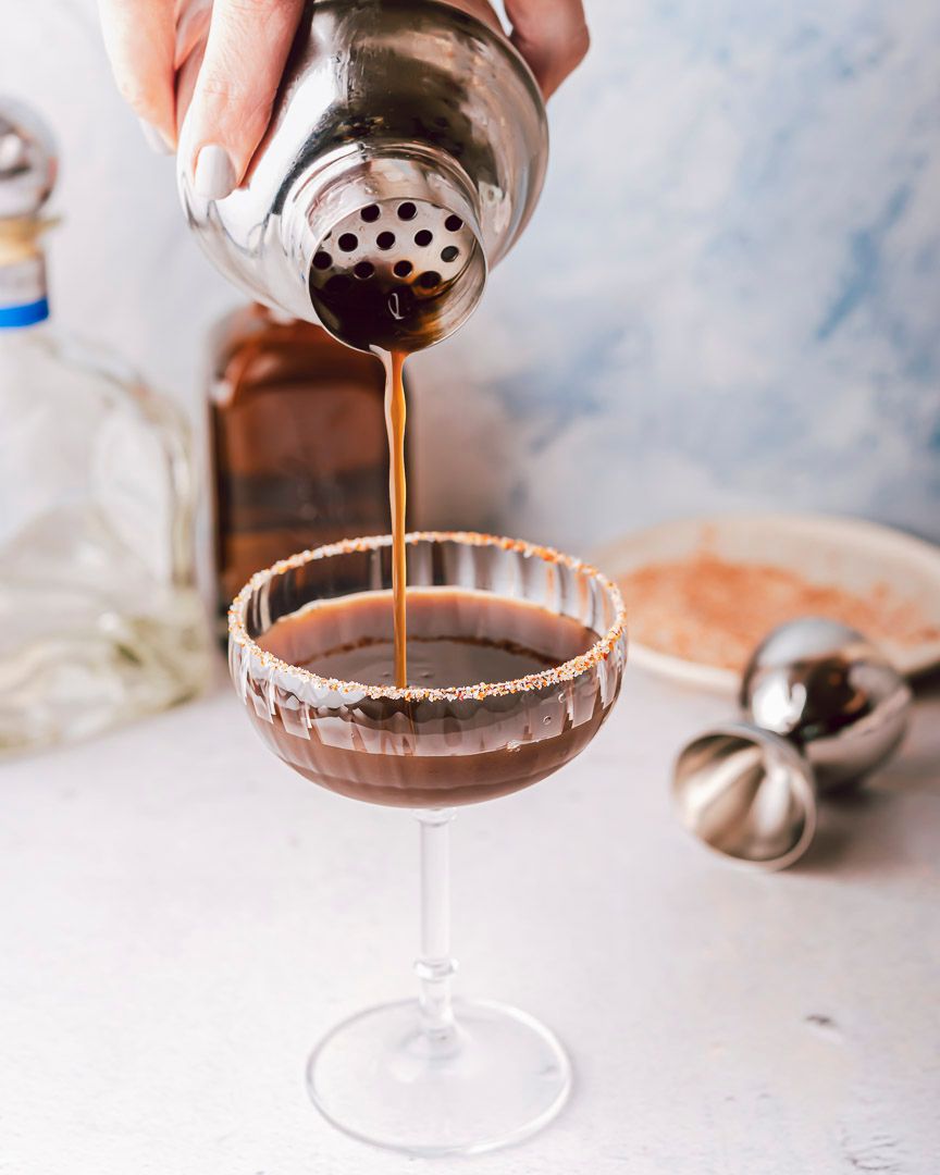 A cocktail shaker pouring a dark brown tamarind cocktail into a coupe glass with a tajin rim.
