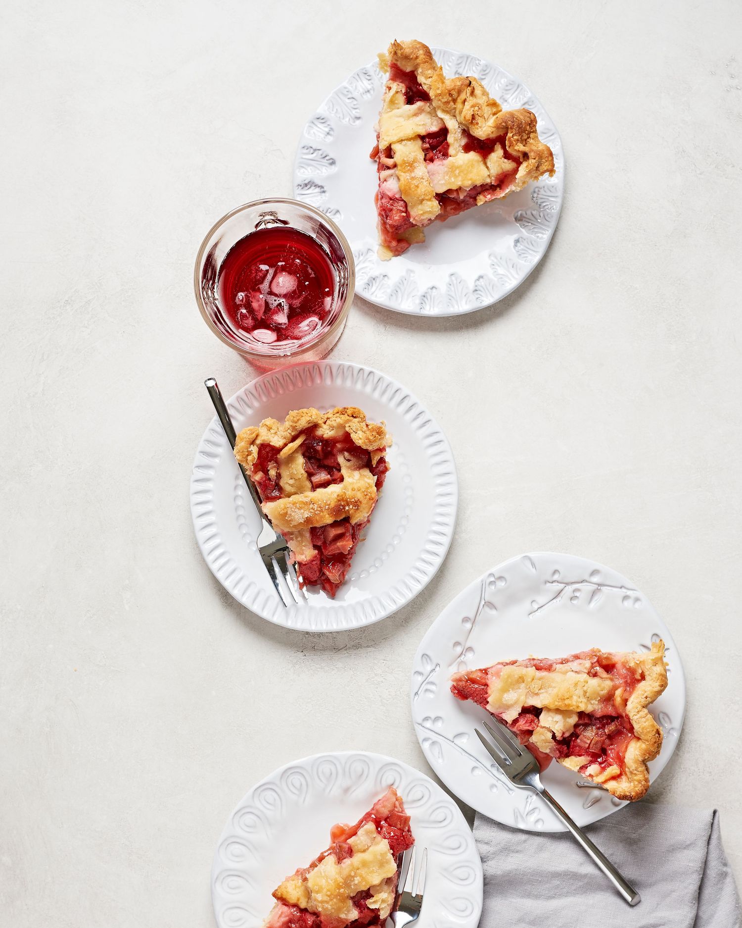 Slices of strawberry rhubarb pie with lattice crust served on white plates with a refreshing hibiscus drink.