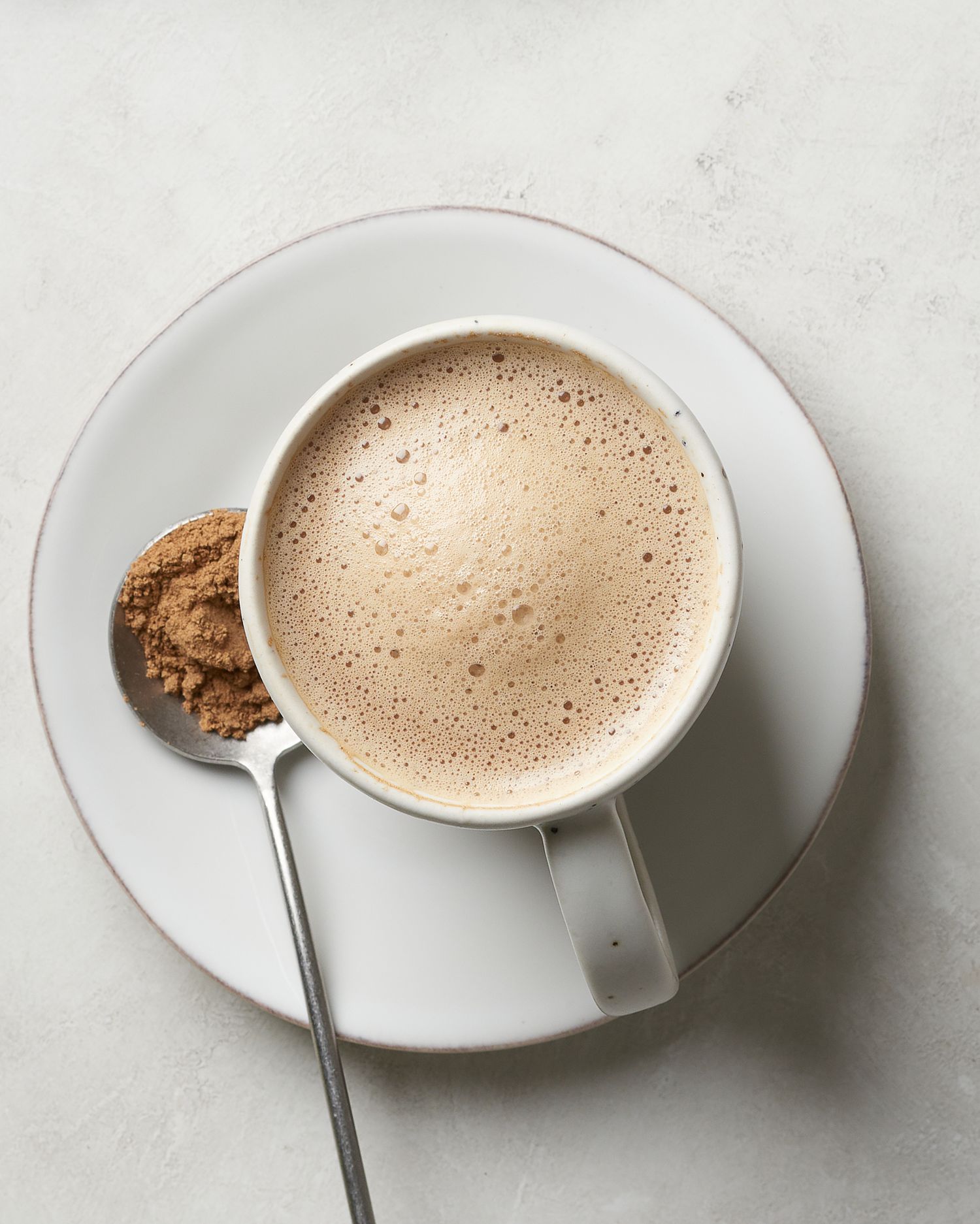 Frothy mushroom latte in a white cup with mushroom powder on a saucer against a light background.