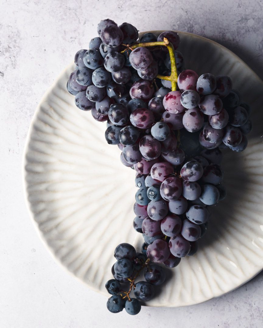 Fresh purple grape cluster artfully arranged on white ceramic plate.