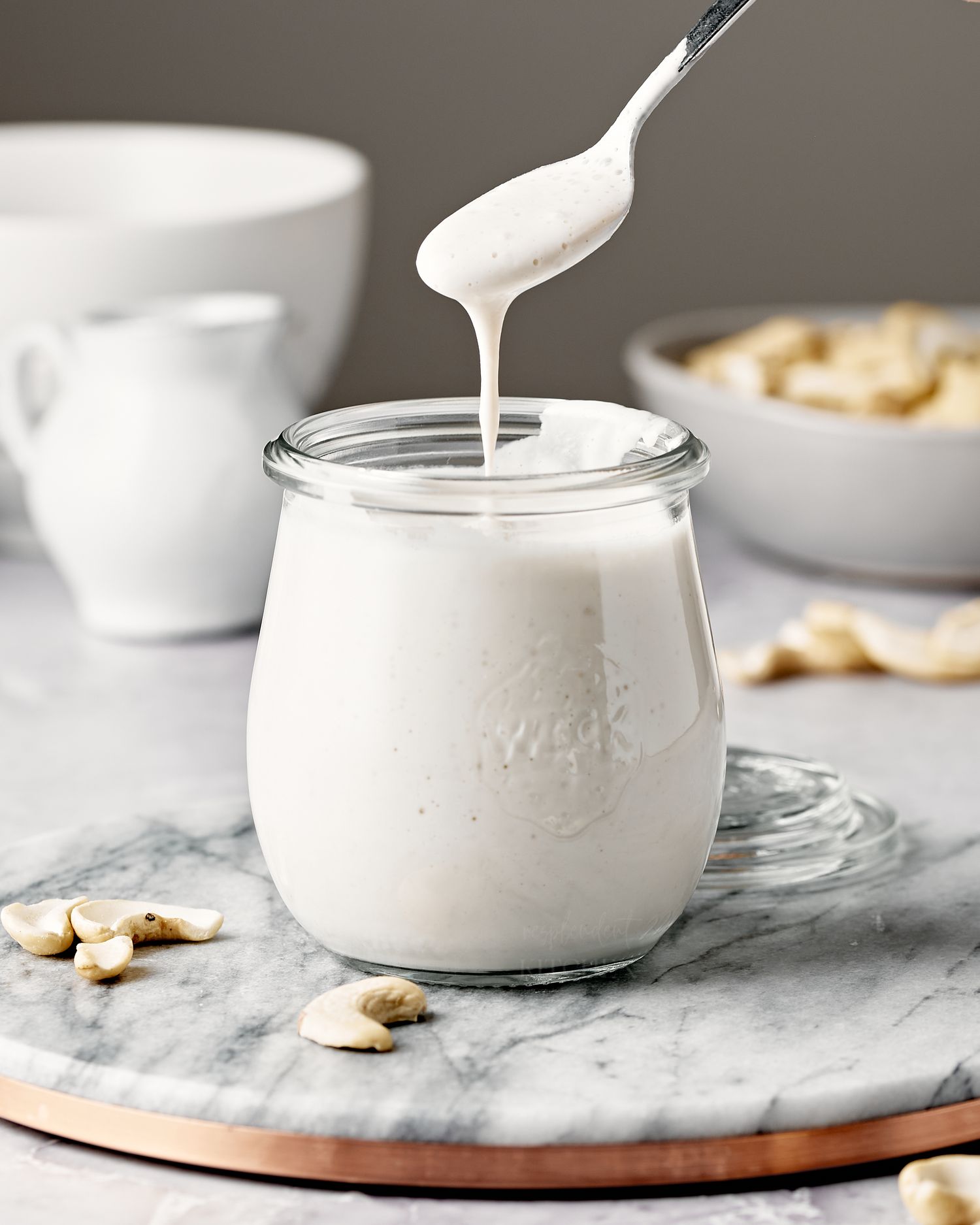 Creamy white cashew cream being spooned from a glass jar on a marble surface with scattered nuts.