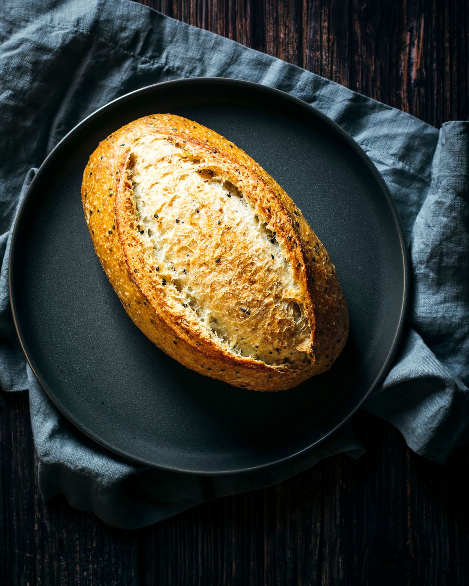 Artisanal bread loaf with crispy crust on black plate with blue linen napkin.