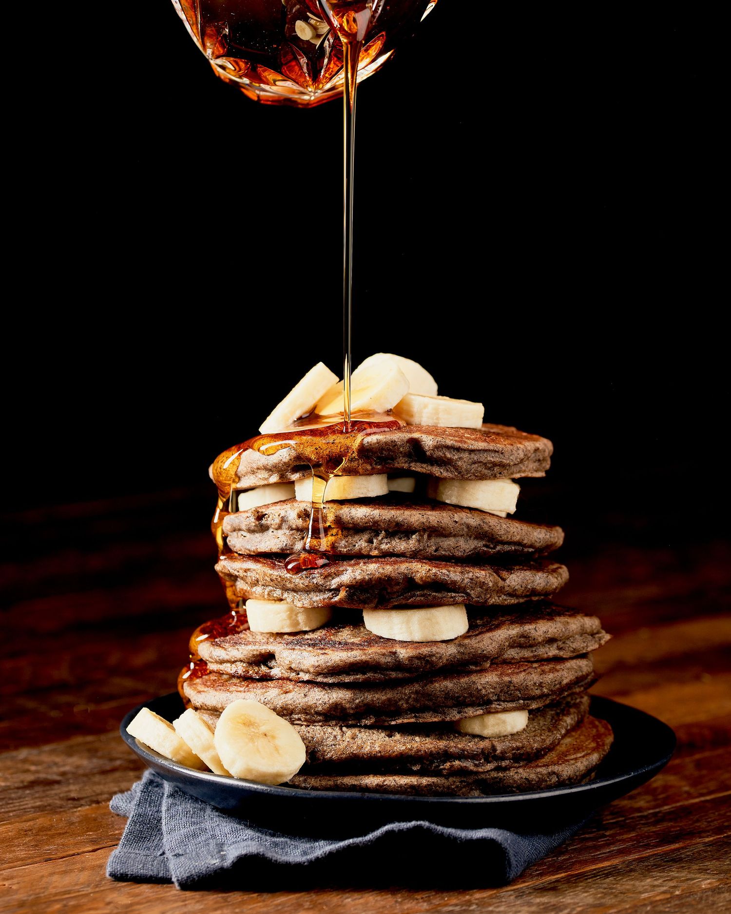 Stack of fluffy pancakes with banana slices and maple syrup drizzle on dark background.