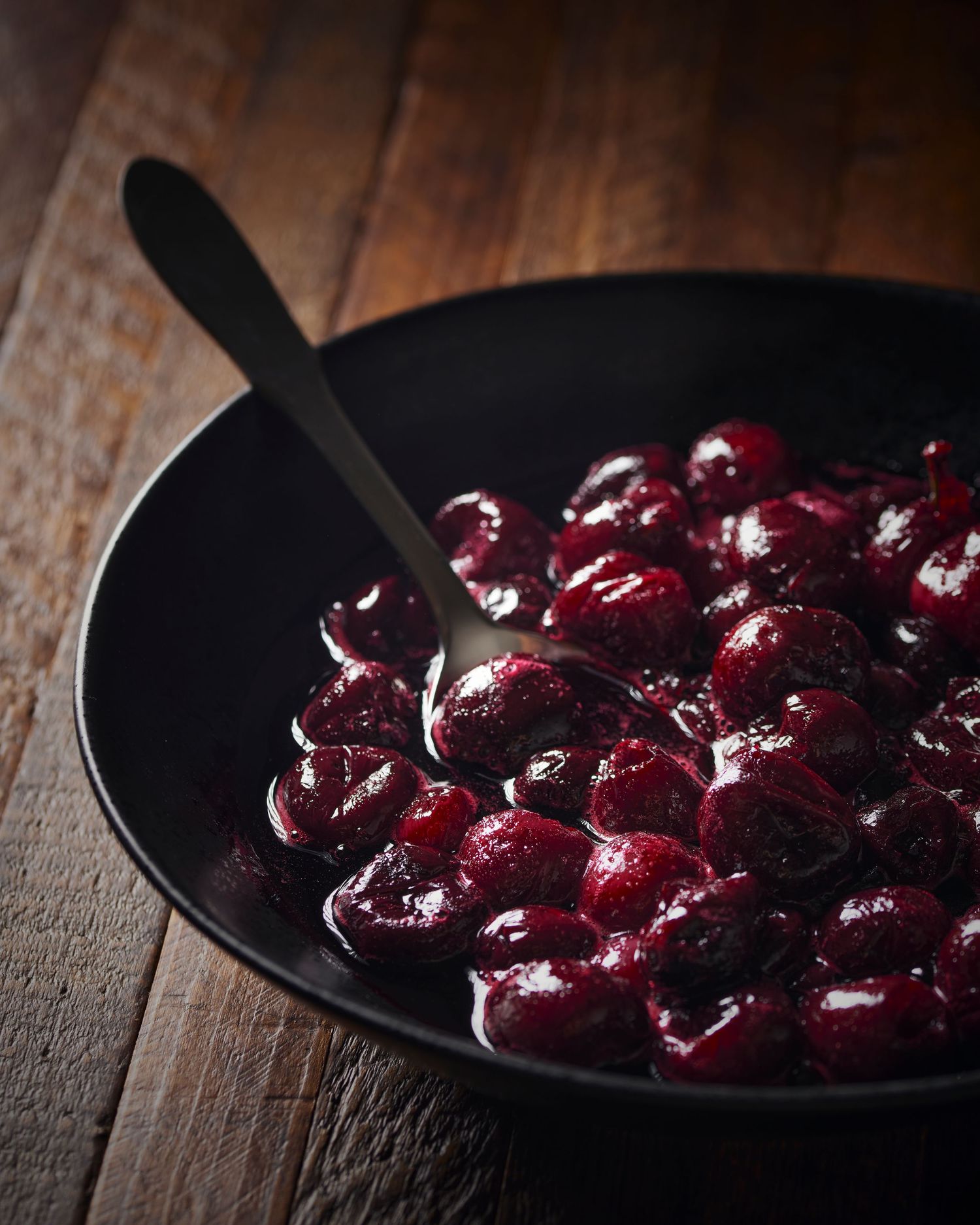 Fresh dark cherries glistening in a black bowl against rustic wooden background.