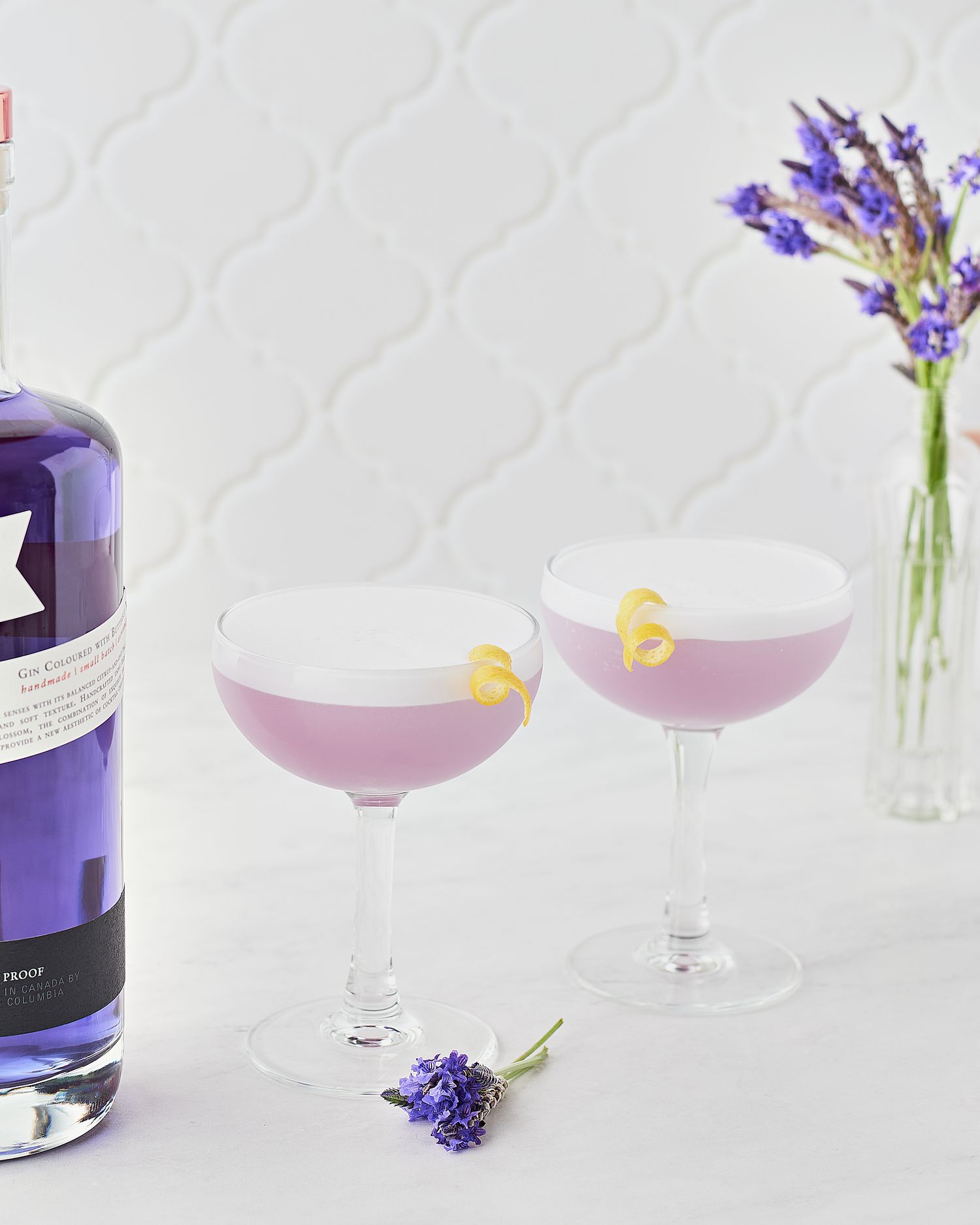 Purple cocktails in coupe glasses with lavender garnish and a bottle on white background.