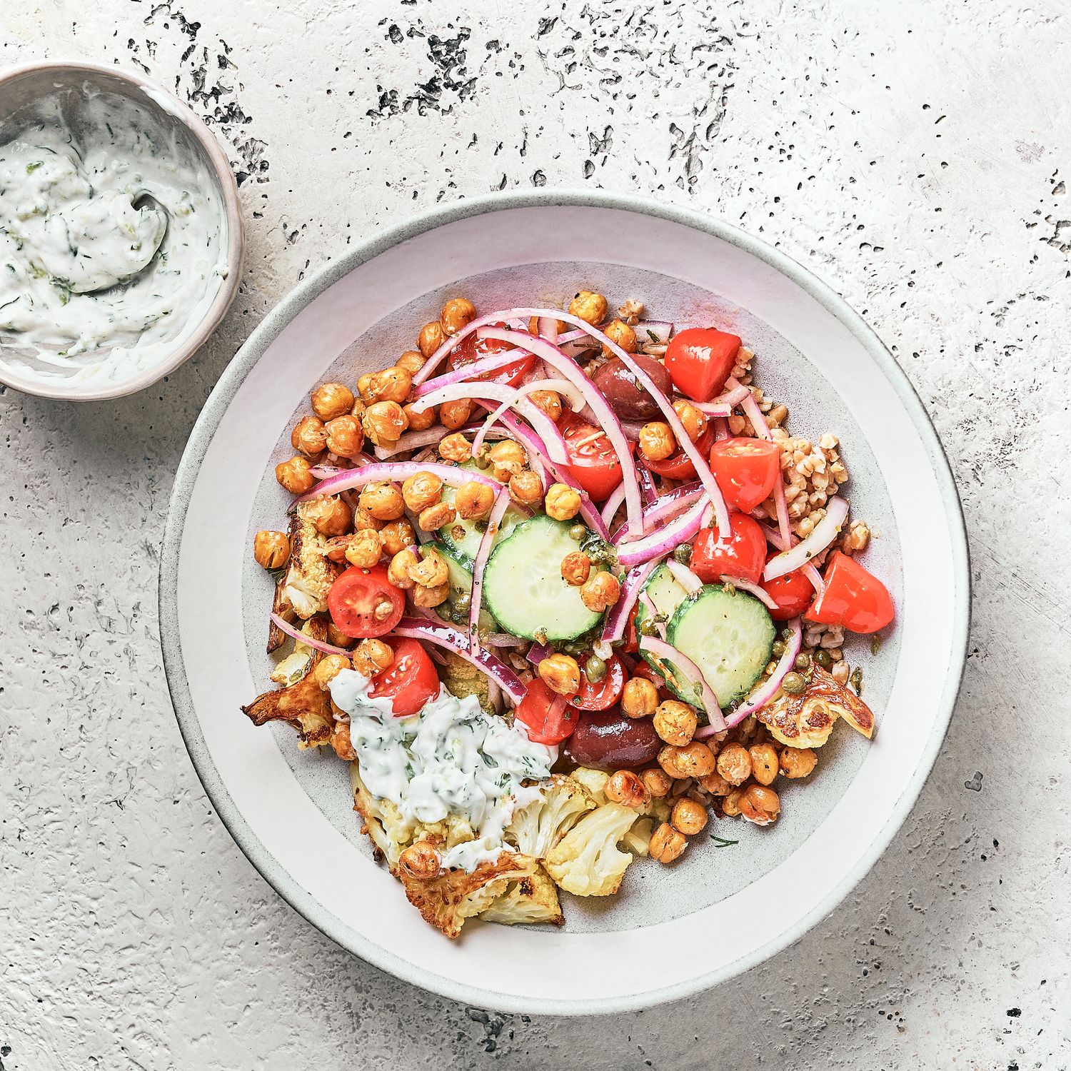 Mediterranean bowl with chickpeas, vegetables, and vegan tzatziki sauce on white plate.