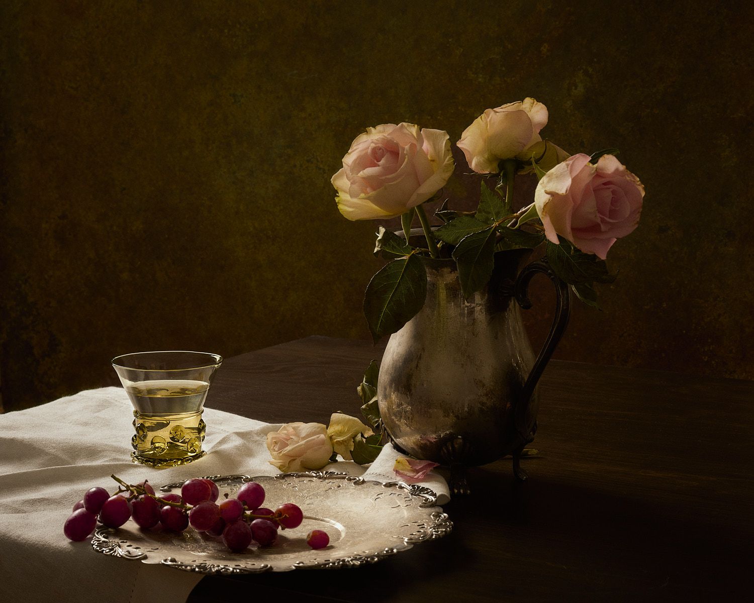 Classic still life with pink roses in silver pitcher, red grapes on tray, and glass of wine on white cloth.