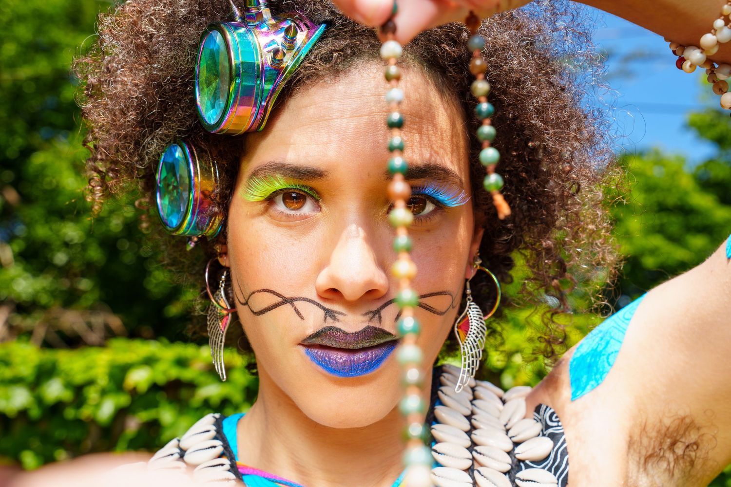 Portrait of a performer in a full colorful costume that includes futuristic steampunk goggles, rosary beads, green & blue eyelashes, jewels