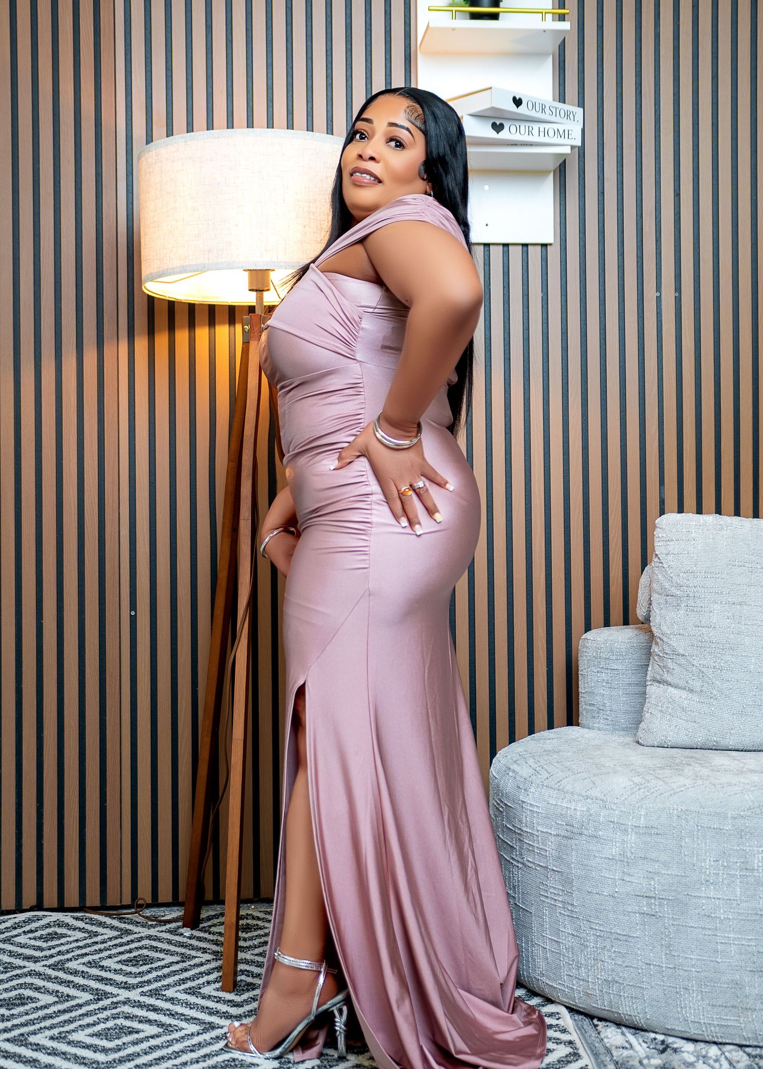 Woman in elegant mauve satin gown with side slit posing next to a floor lamp in a modern interior.