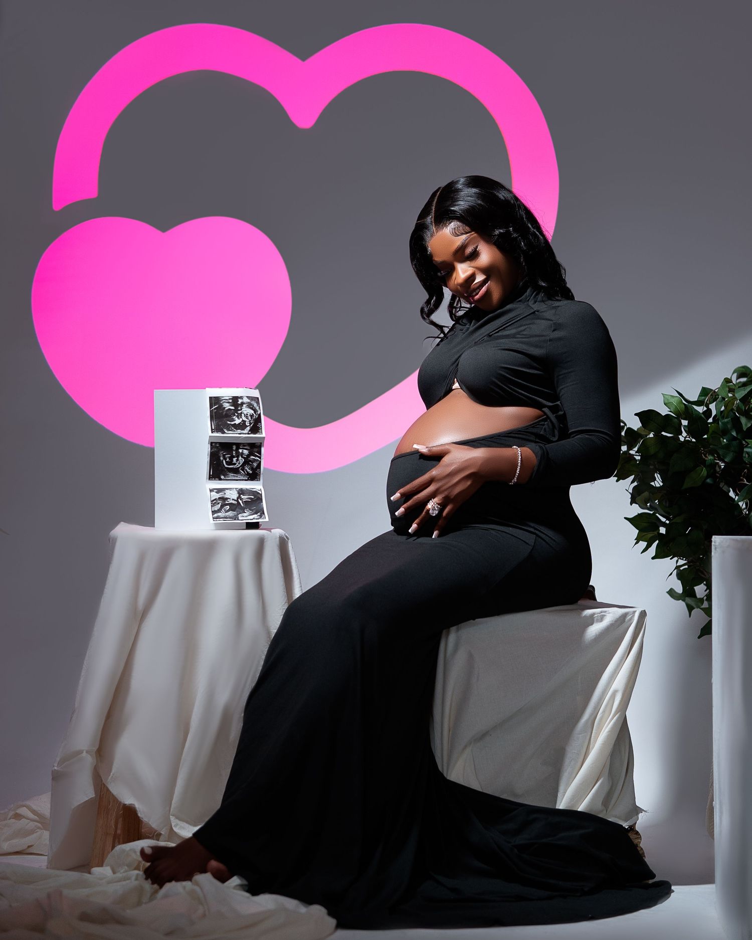 Pregnant woman in black dress poses for maternity photoshoot with ultrasound photos and pink heart backdrop.