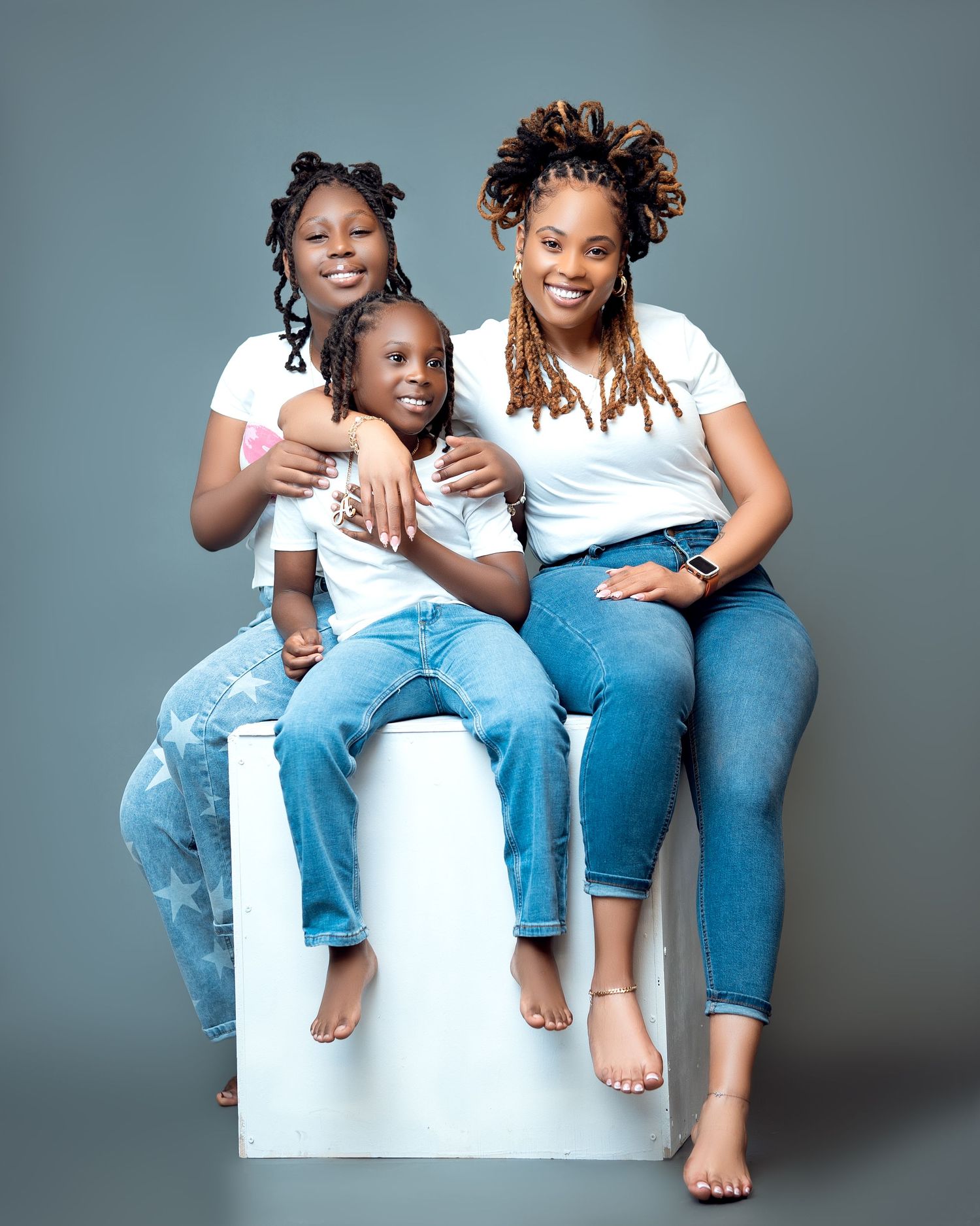 A woman and two young girls in matching white tops and jeans pose together for a studio portrait photo.