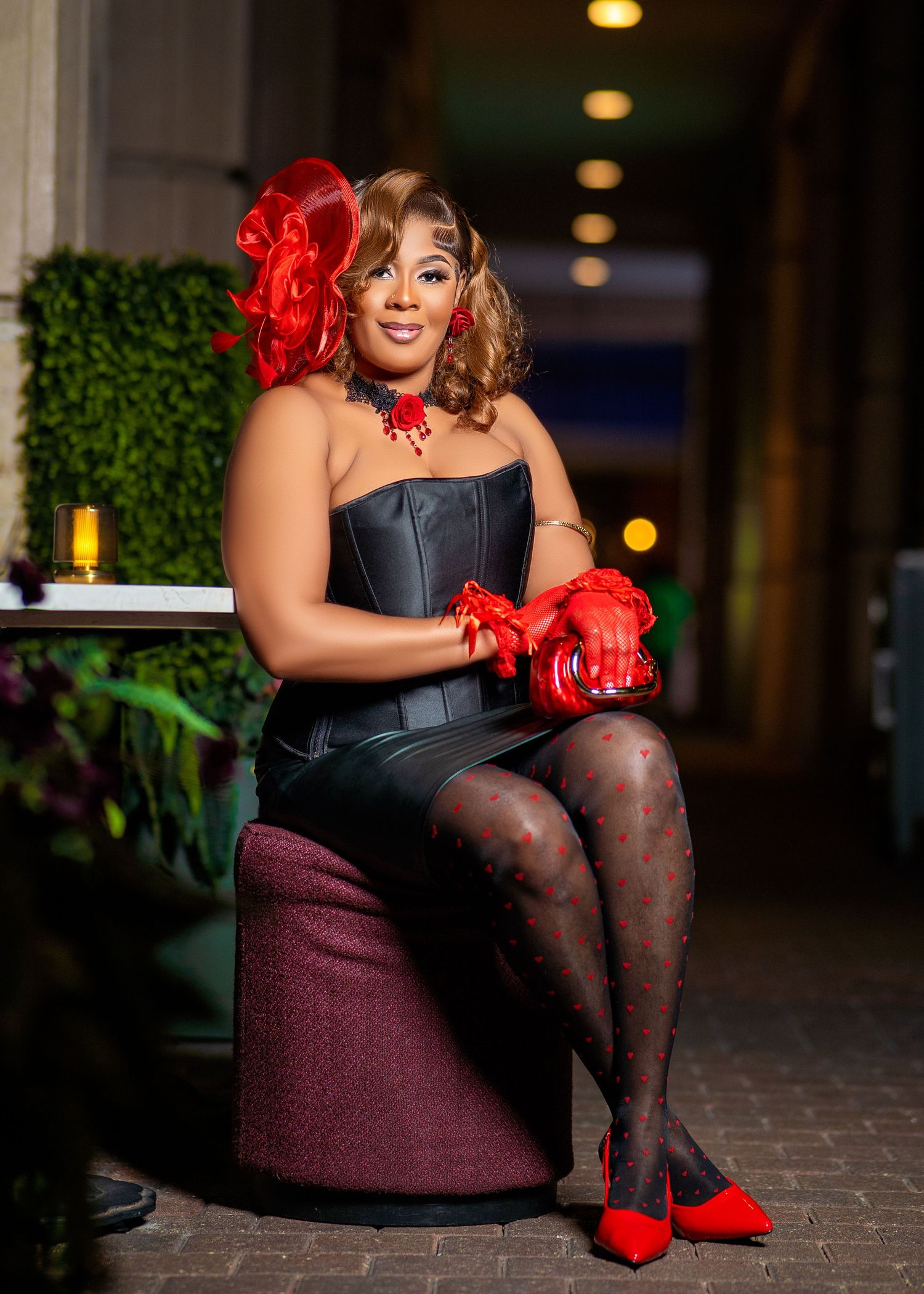 Woman in black corset and red accessories poses elegantly on dark stool.