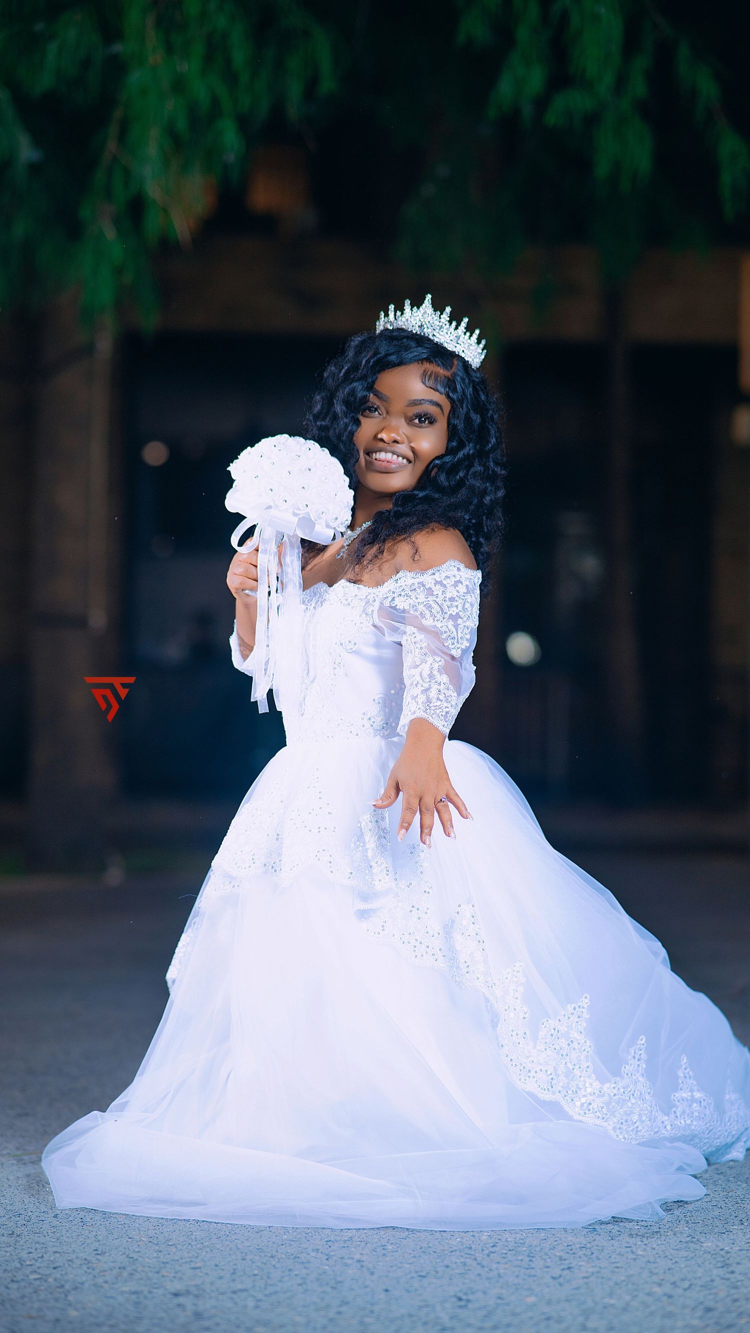 A person in an elegant white lace wedding gown poses gracefully on the ground with a tiara.