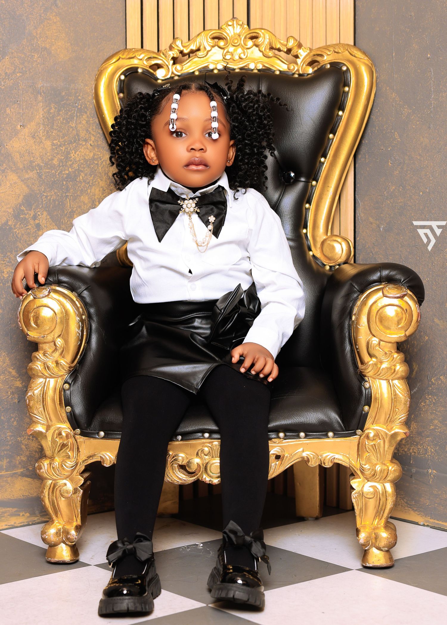 Child in formal black and white outfit sits on ornate gold throne chair.