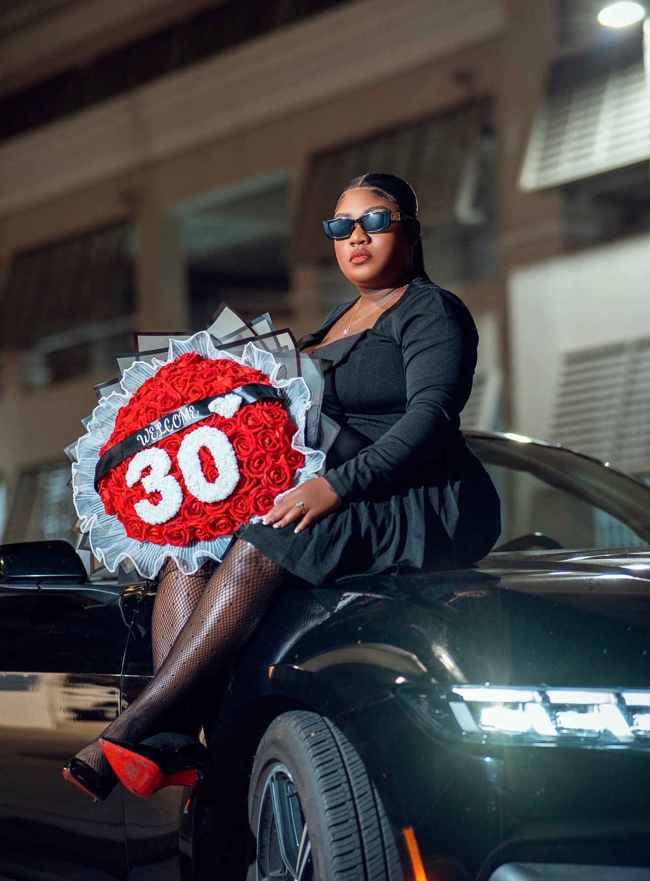 Woman in black dress celebrating 30th birthday, posing with red rose bouquet on luxury car at night.
