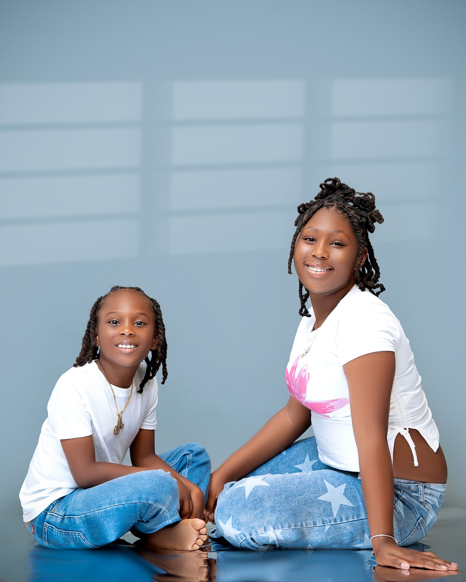 Two young girls in white tops and blue jeans sitting cross-legged on a blue floor against a gray background.