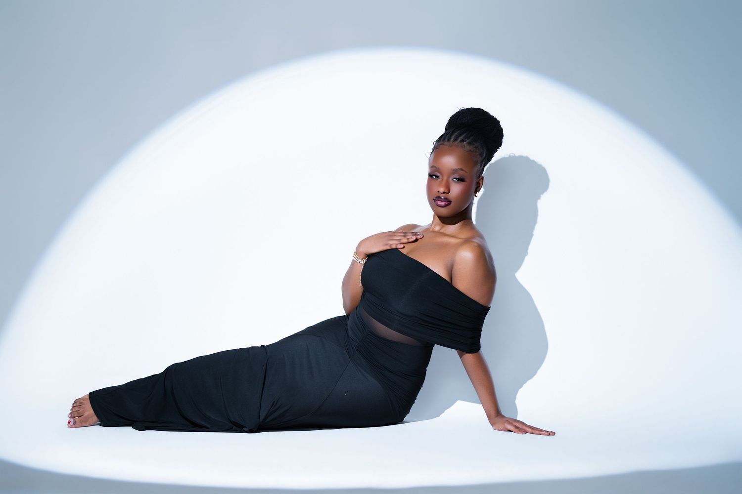 A woman in a black off-shoulder outfit poses gracefully on a white studio floor under a circular spotlight.