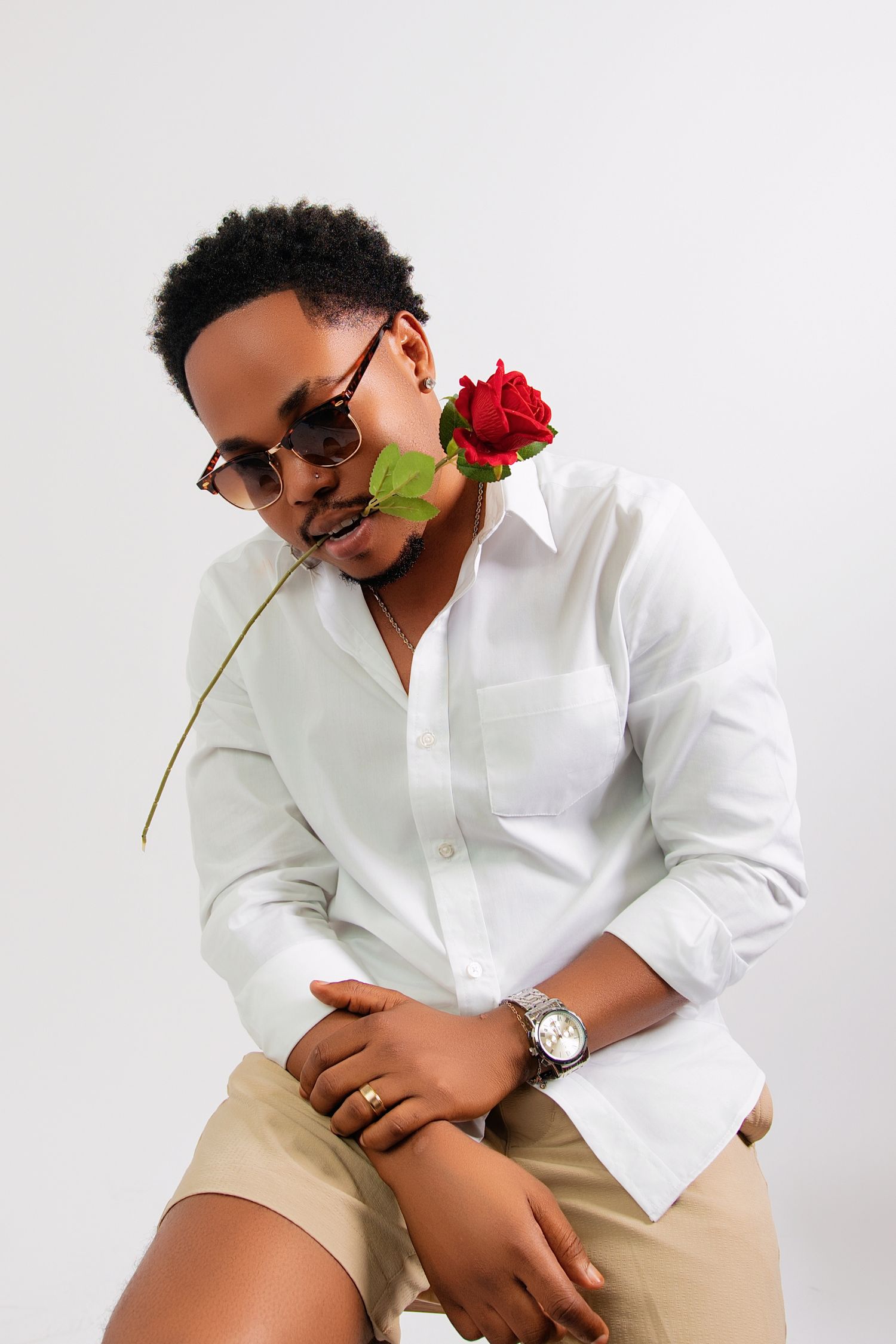 Stylish young Black man in white shirt and sunglasses holding a red rose in his mouth while seated against white background.