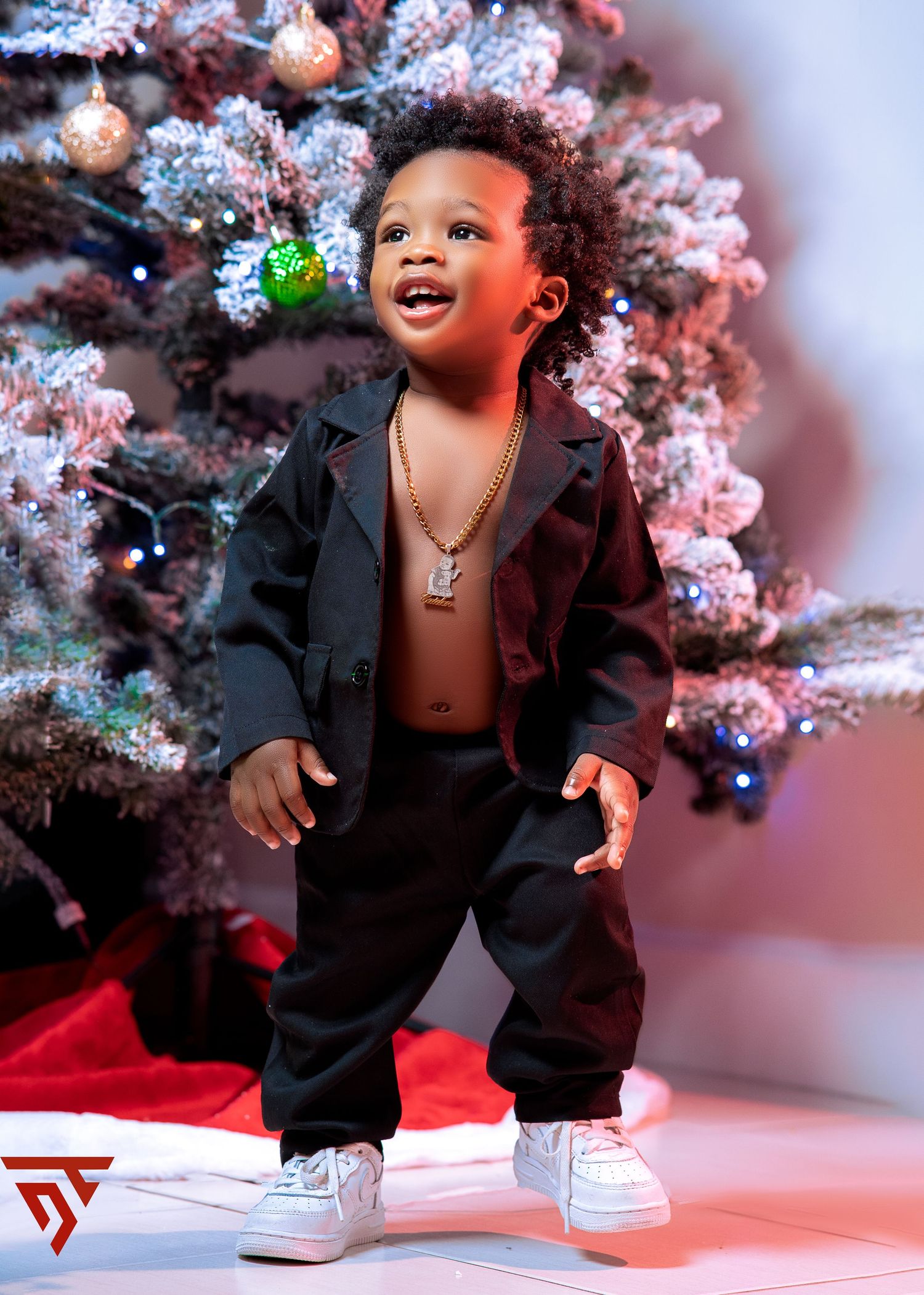 A festive holiday photoshoot featuring a black suit and sneakers against a snowy Christmas tree backdrop.