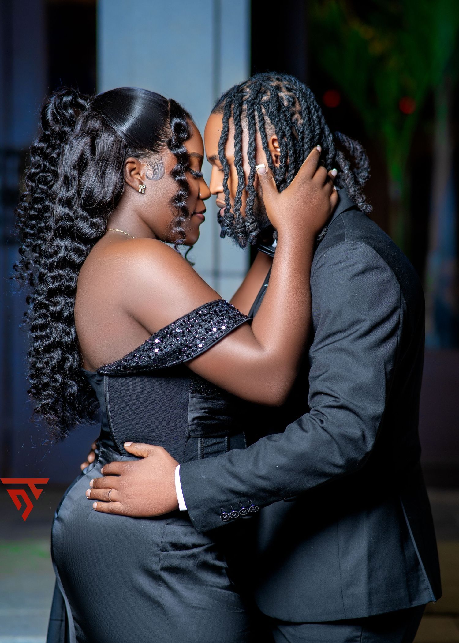 A couple in formal black attire embrace in a romantic outdoor photoshoot.