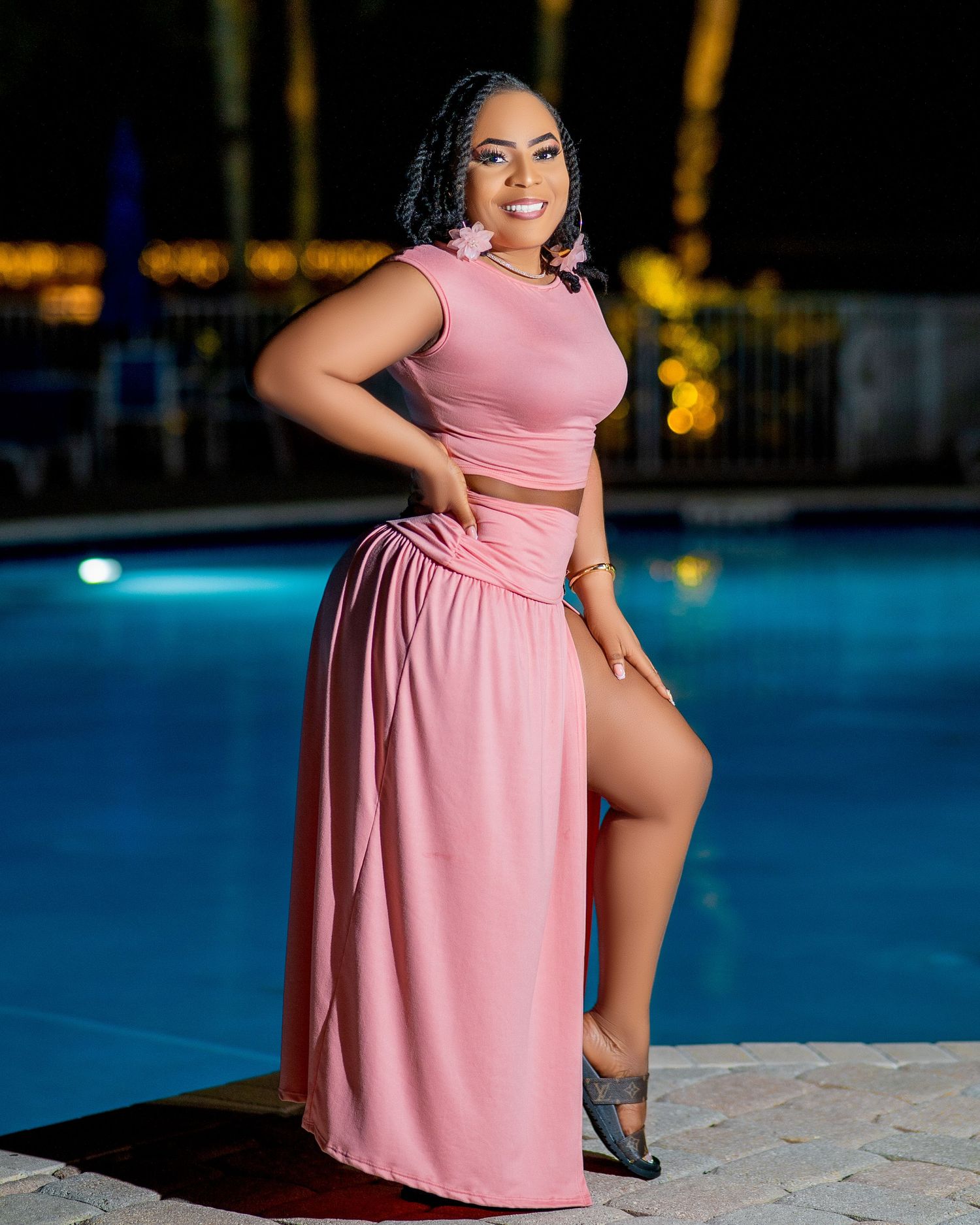 Woman in pink two-piece dress with slit poses confidently by illuminated pool at night.