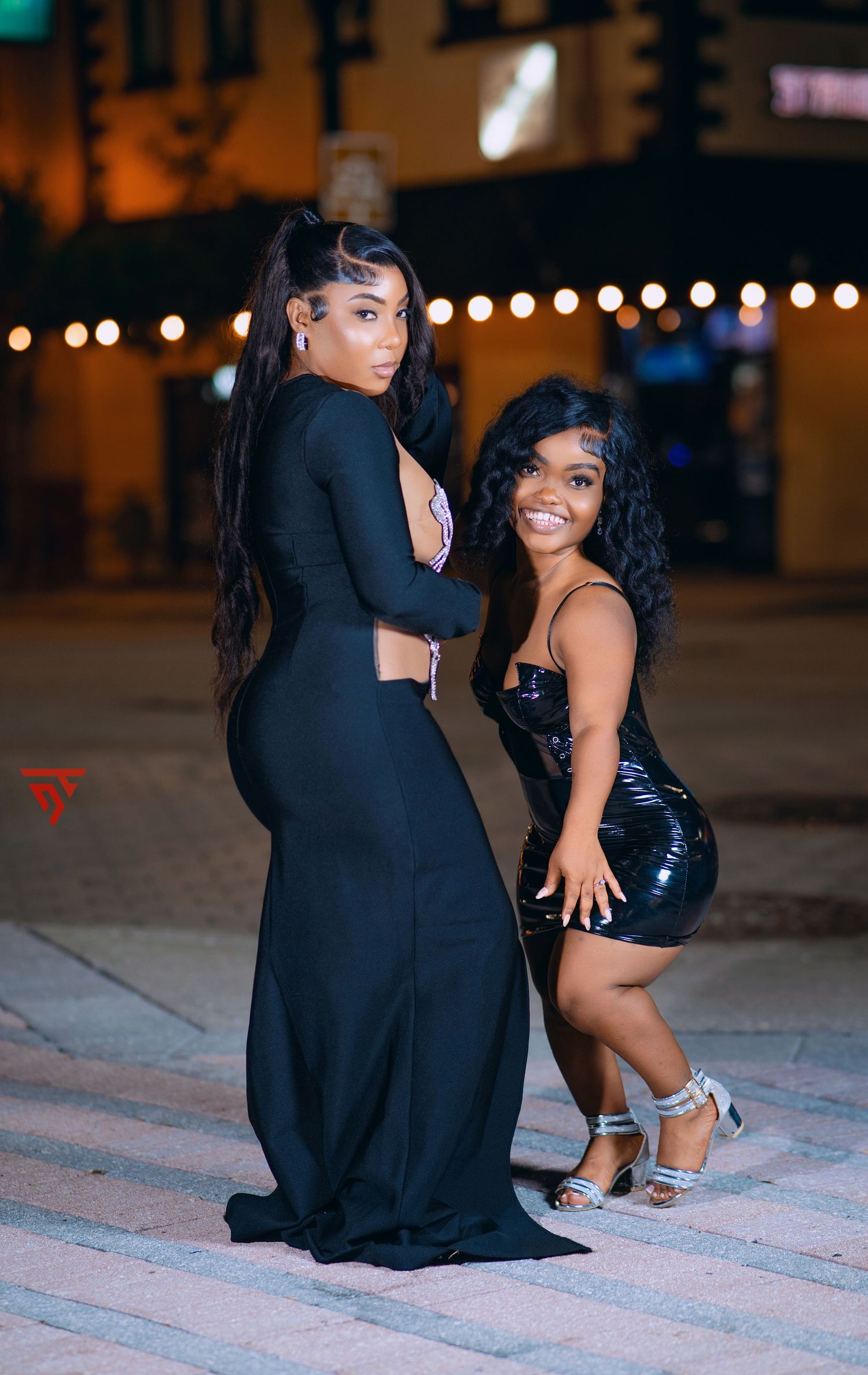 Two friends posing together at night, wearing elegant black dresses against city lights.