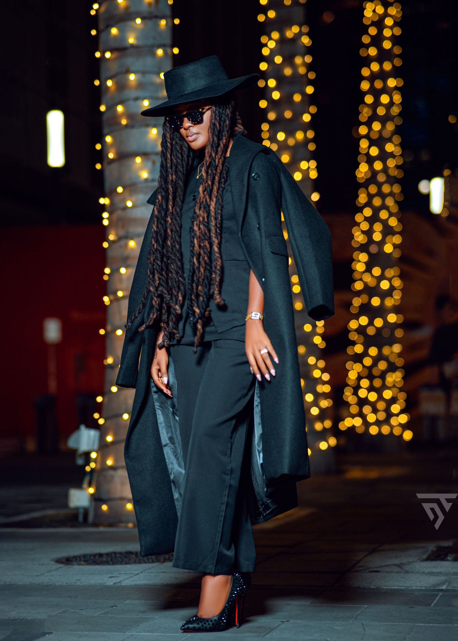 Dramatic nighttime fashion shoot with black outfit, hat and long braids against illuminated background.
