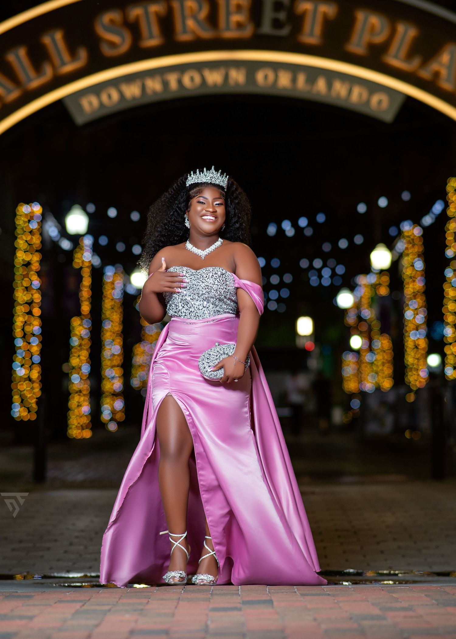 Fashion model in pink formal gown poses under illuminated downtown street sign at night.