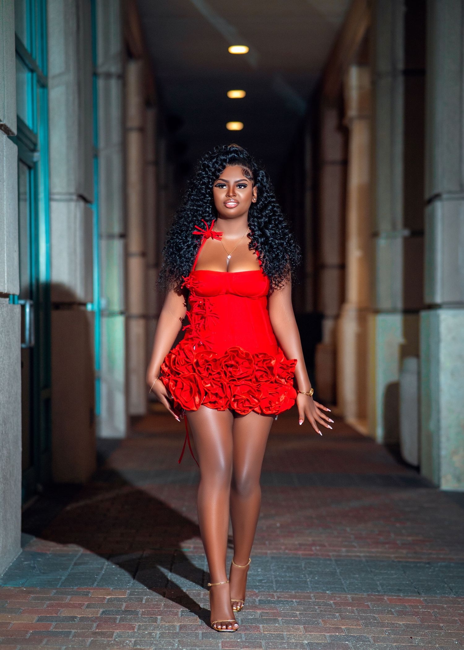Woman in red floral mini dress with curly hair poses confidently in a corridor at night.